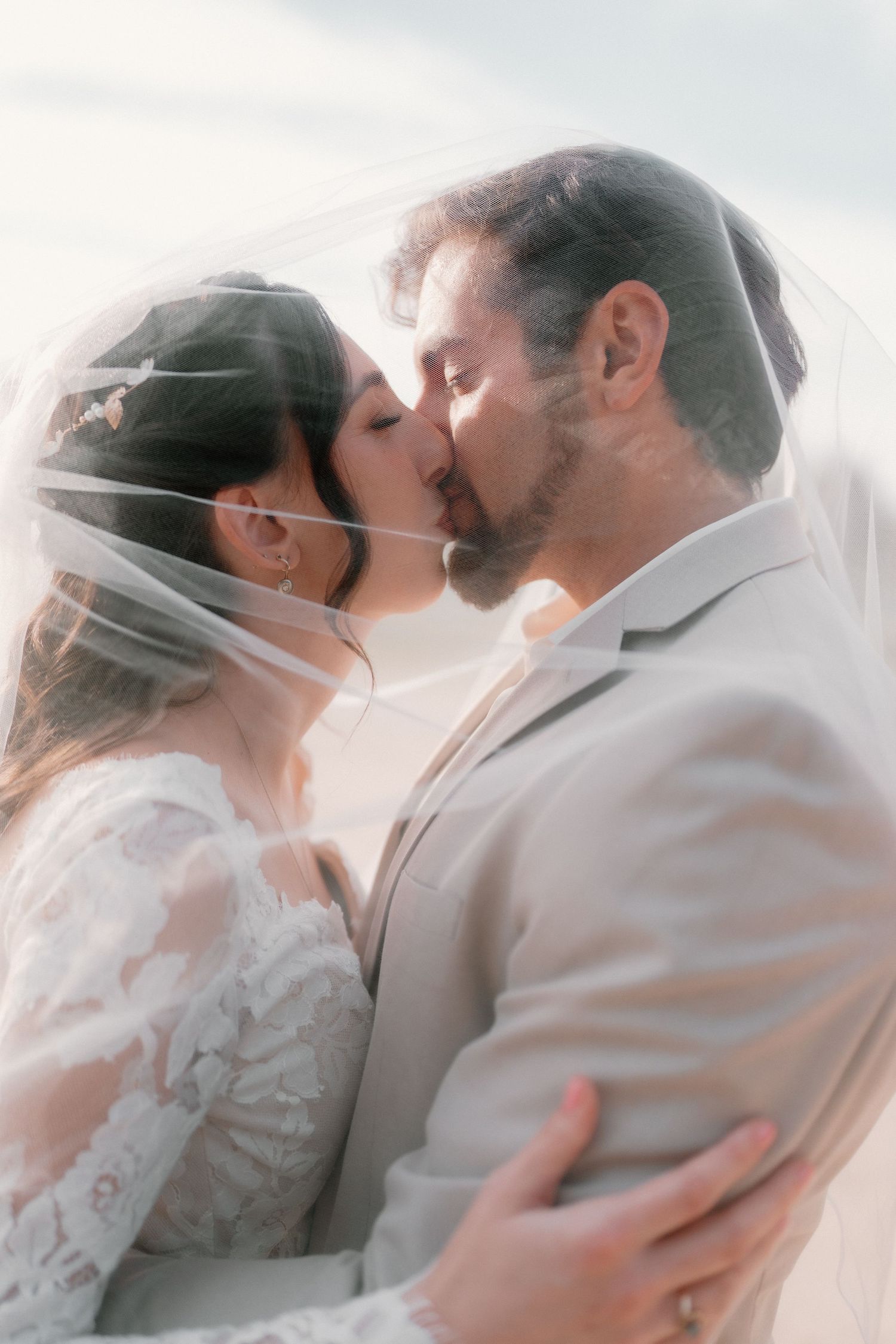 Romantic bride and groom sharing a kiss under sheer wedding veil in soft natural light.