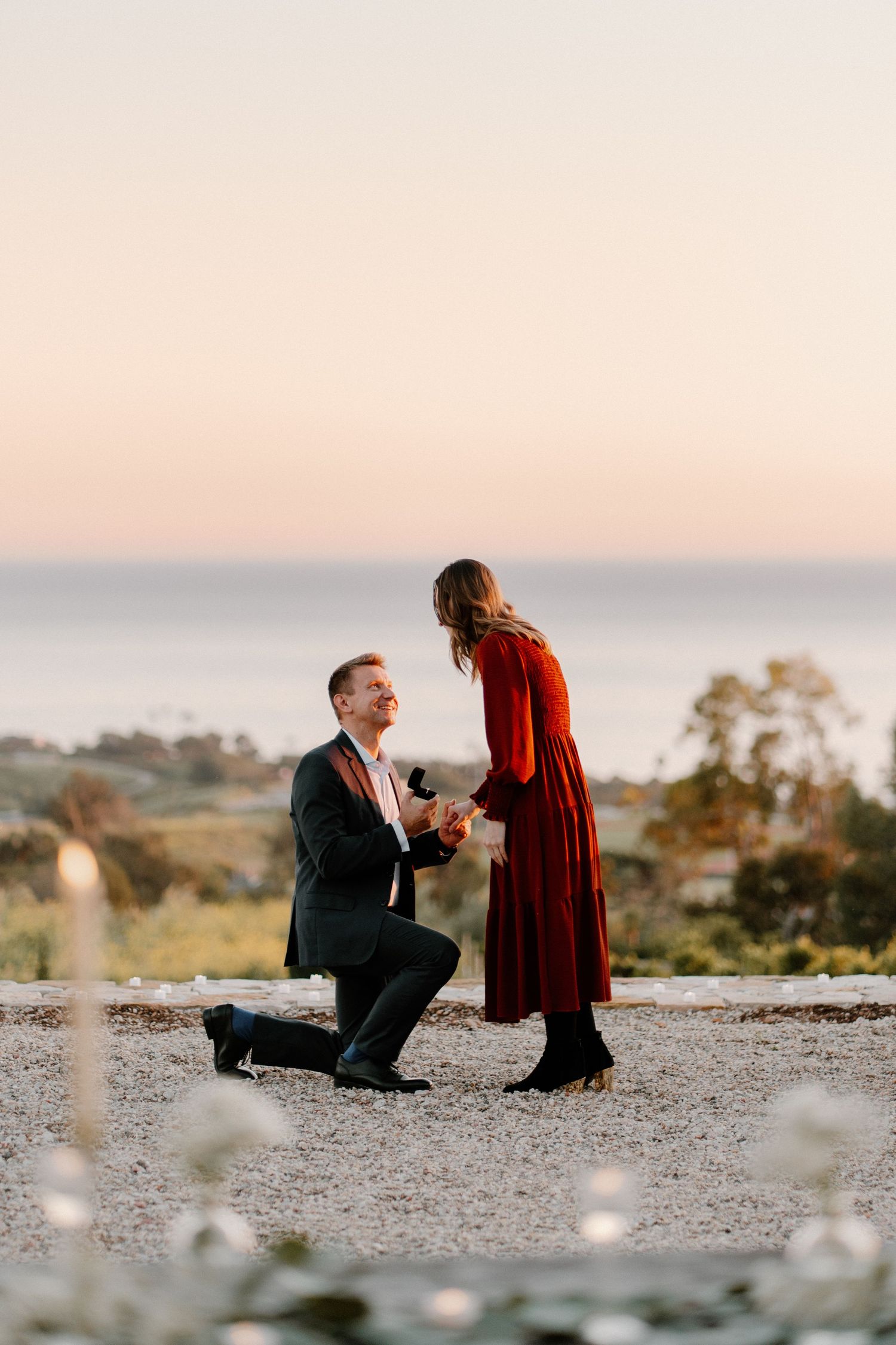 Man proposes on one knee to woman in red dress at sunset by the sea.