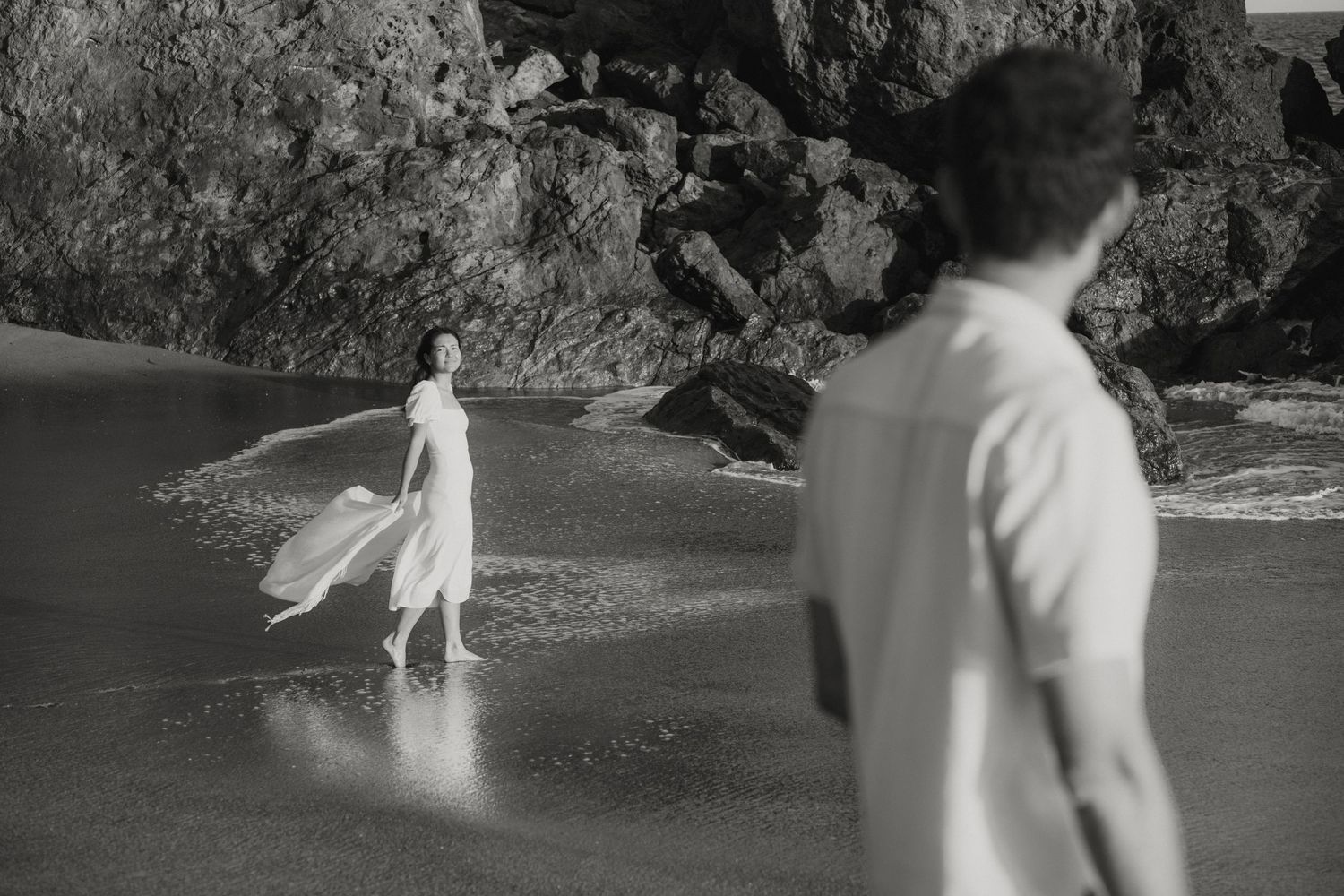 Black and white photo of woman in flowing white dress walking on rocky beach toward man.