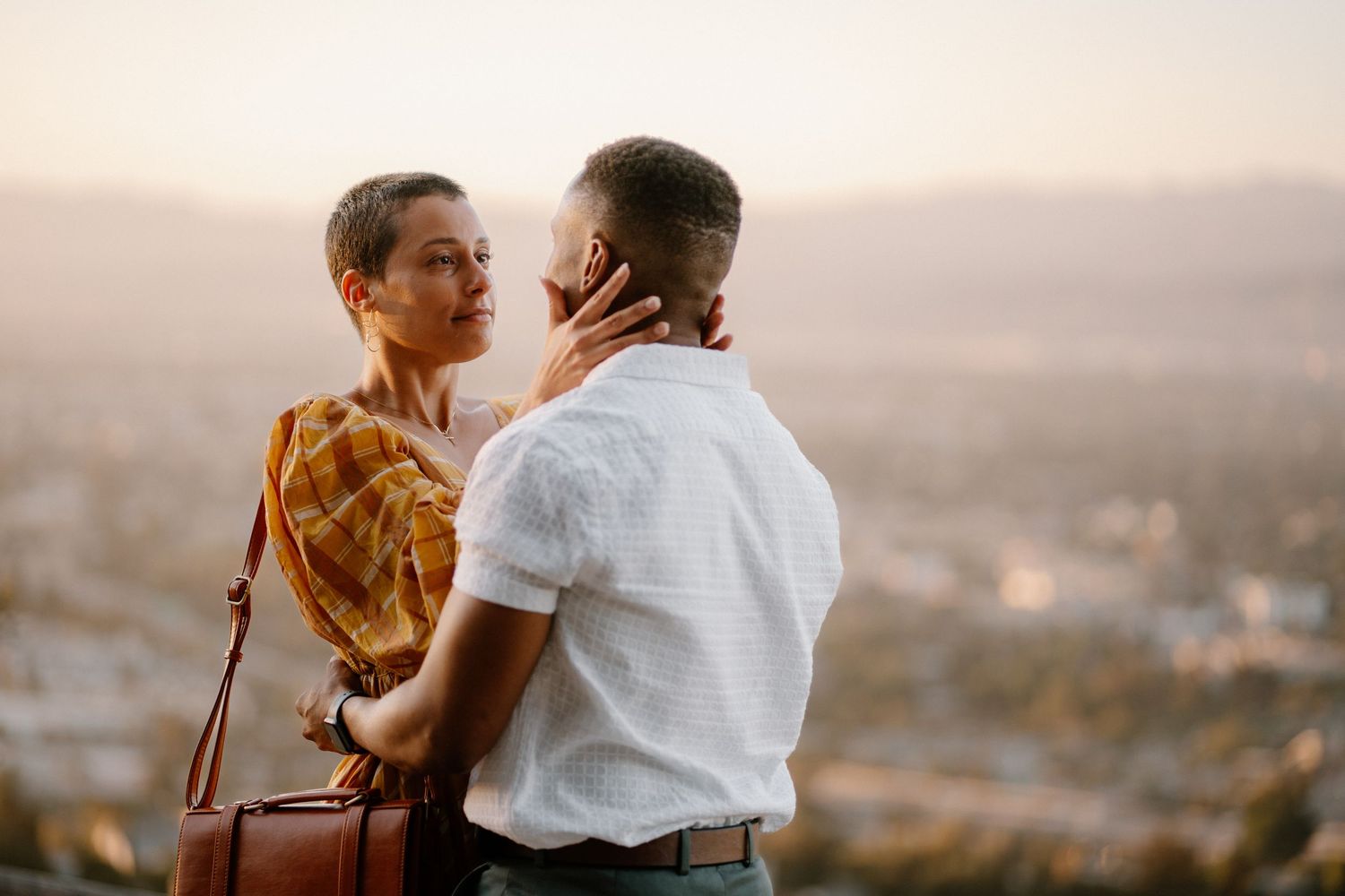 A couple shares a romantic moment at sunset overlooking a city skyline, the woman tenderly holding the man's face.