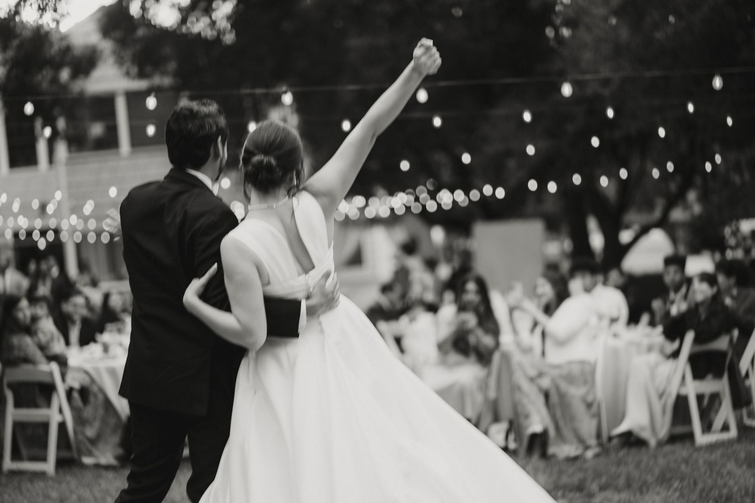 Black and white photo of bride raising fist while dancing with groom at outdoor wedding reception.