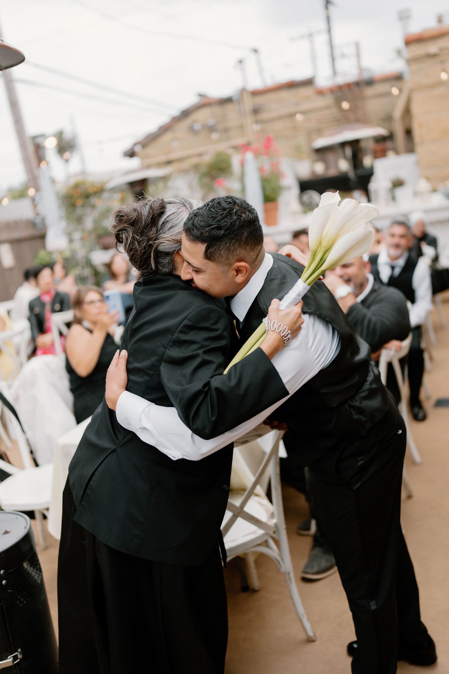 Groom embracing a guest in black at an outdoor wedding reception while holding white calla lilies.