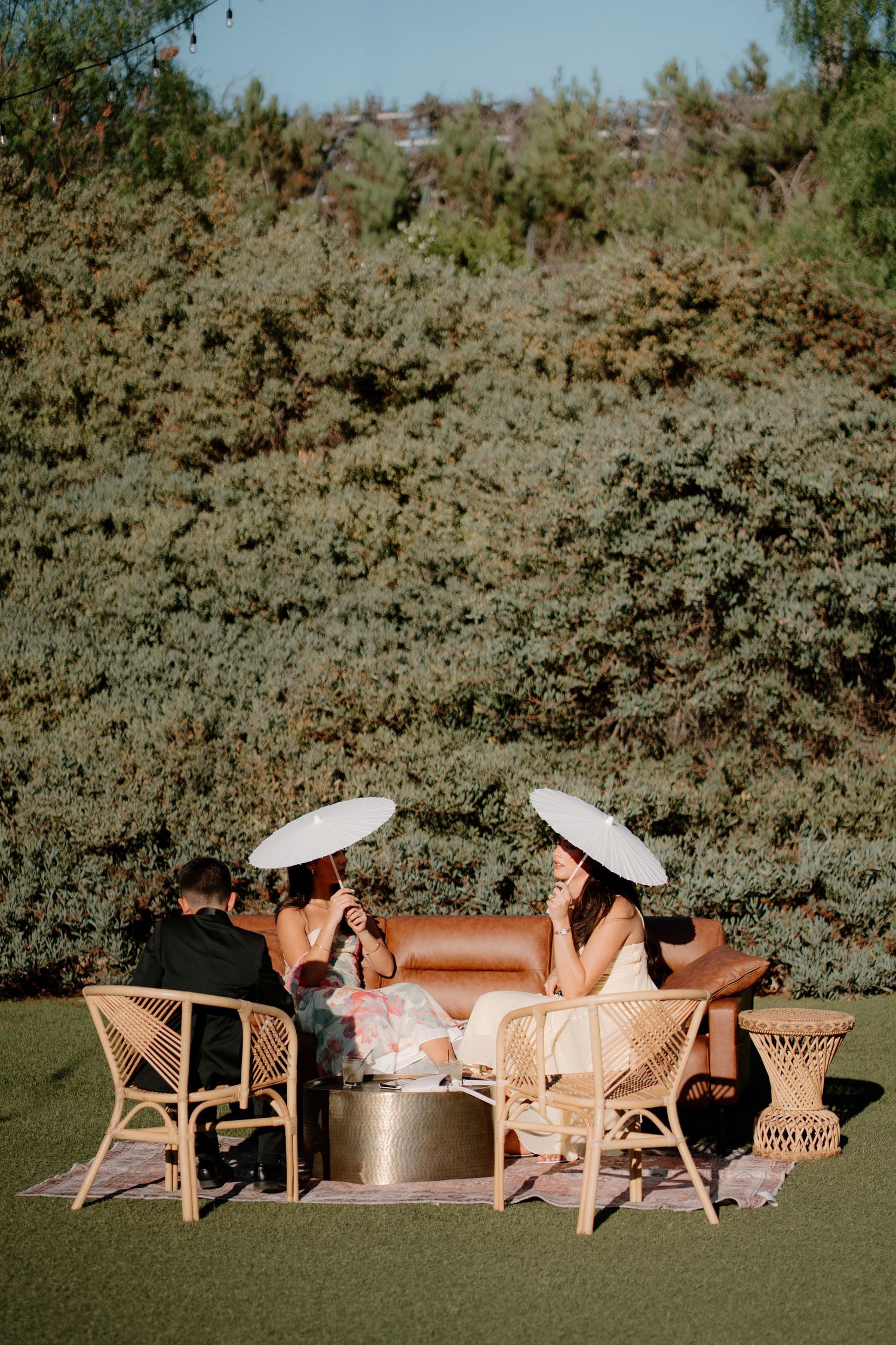 Wedding guests relax on rattan furniture with white parasols in sunlit outdoor garden setting.
