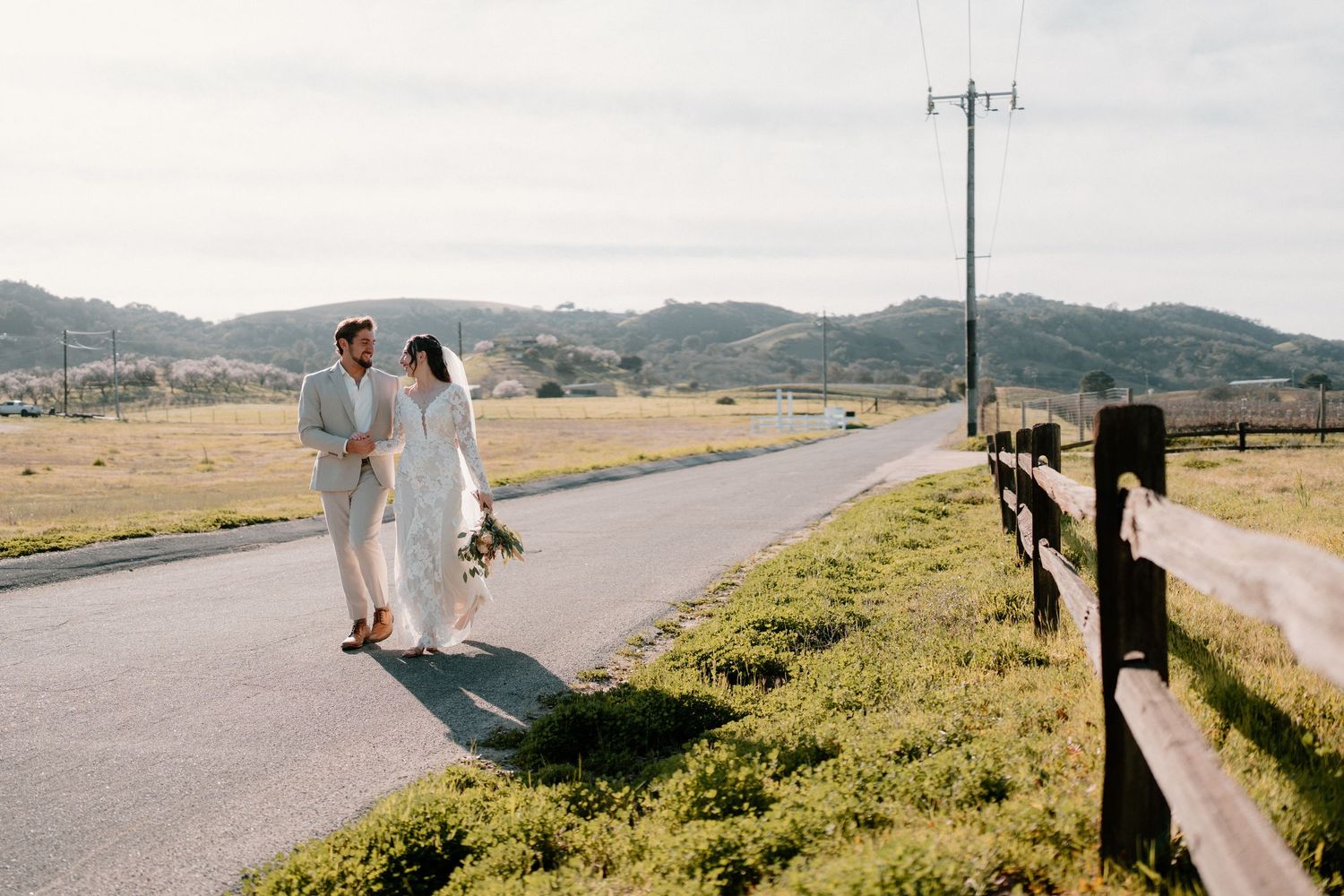 Smiling bride and groom walking together along scenic country road with green hills and fence.