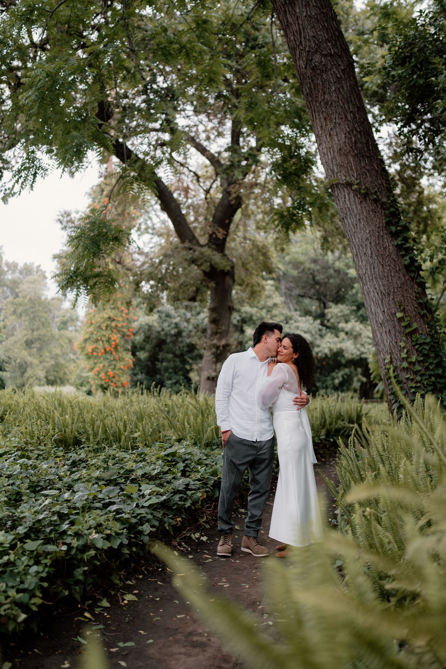 A couple shares a tender embrace on a garden path surrounded by lush green hedges and tall trees.