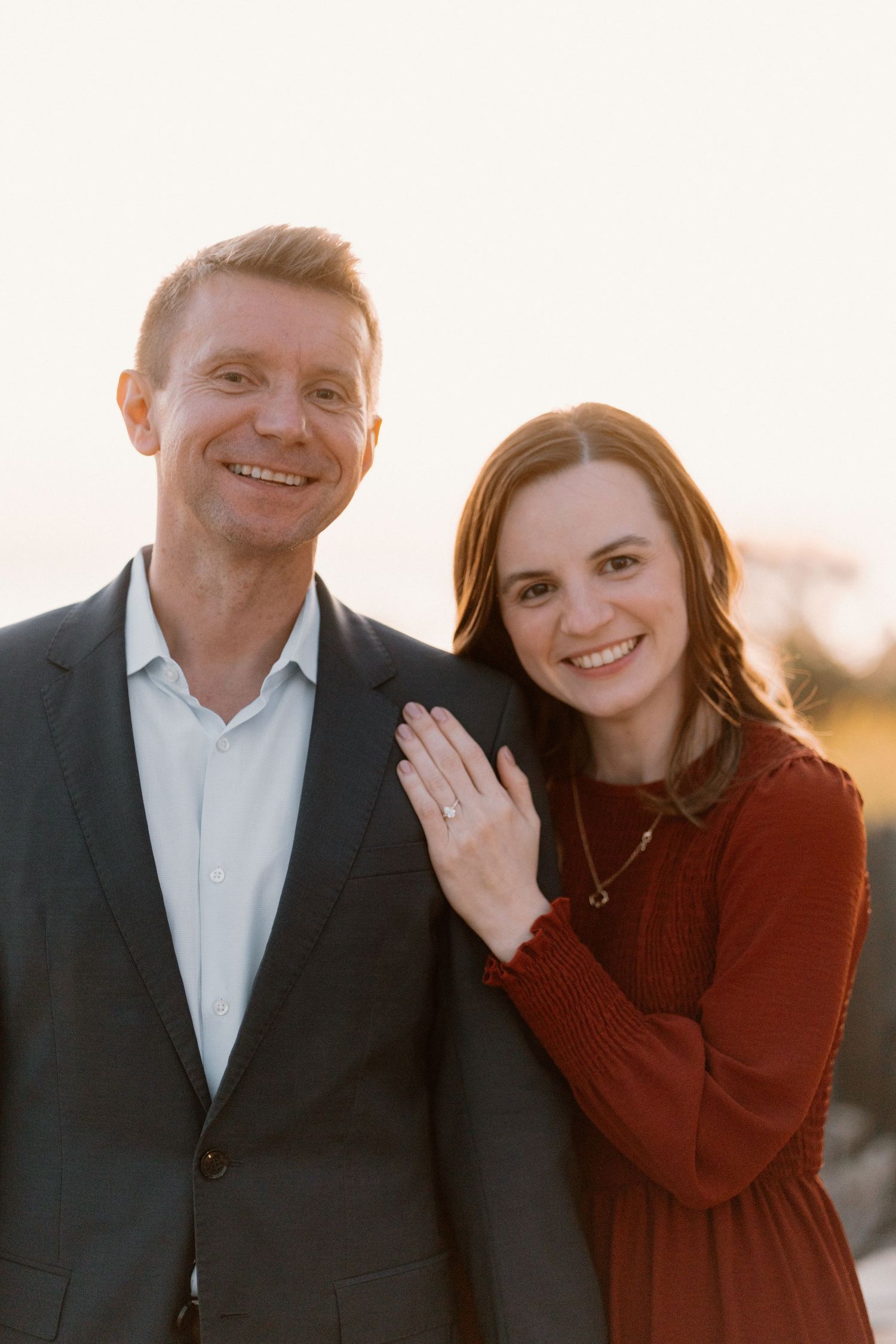 Smiling engaged couple posing together at sunset, woman in red dress displaying diamond ring.