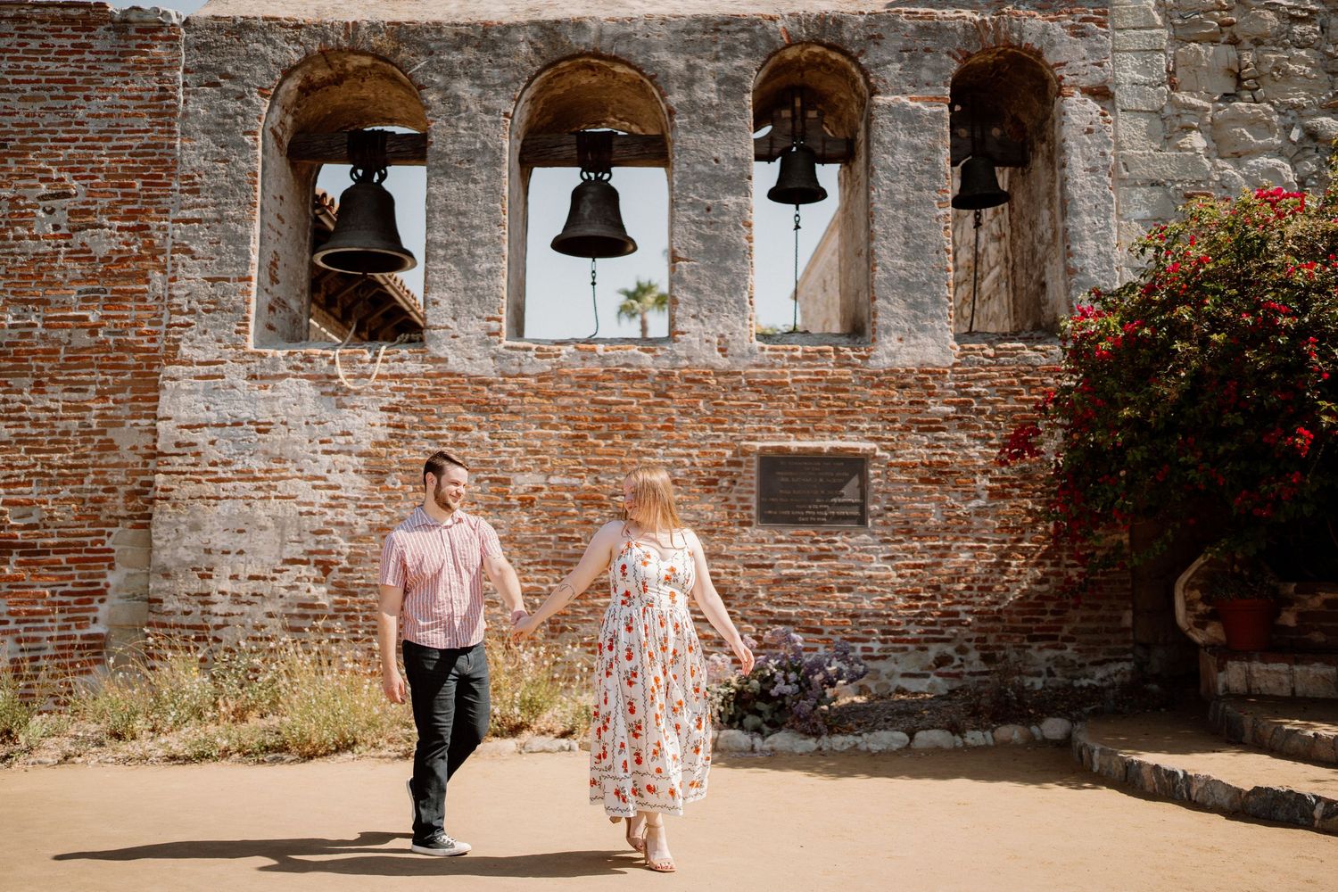Couple holding hands walking in front of historic Mission San Juan Capistrano bell wall with brick arches.