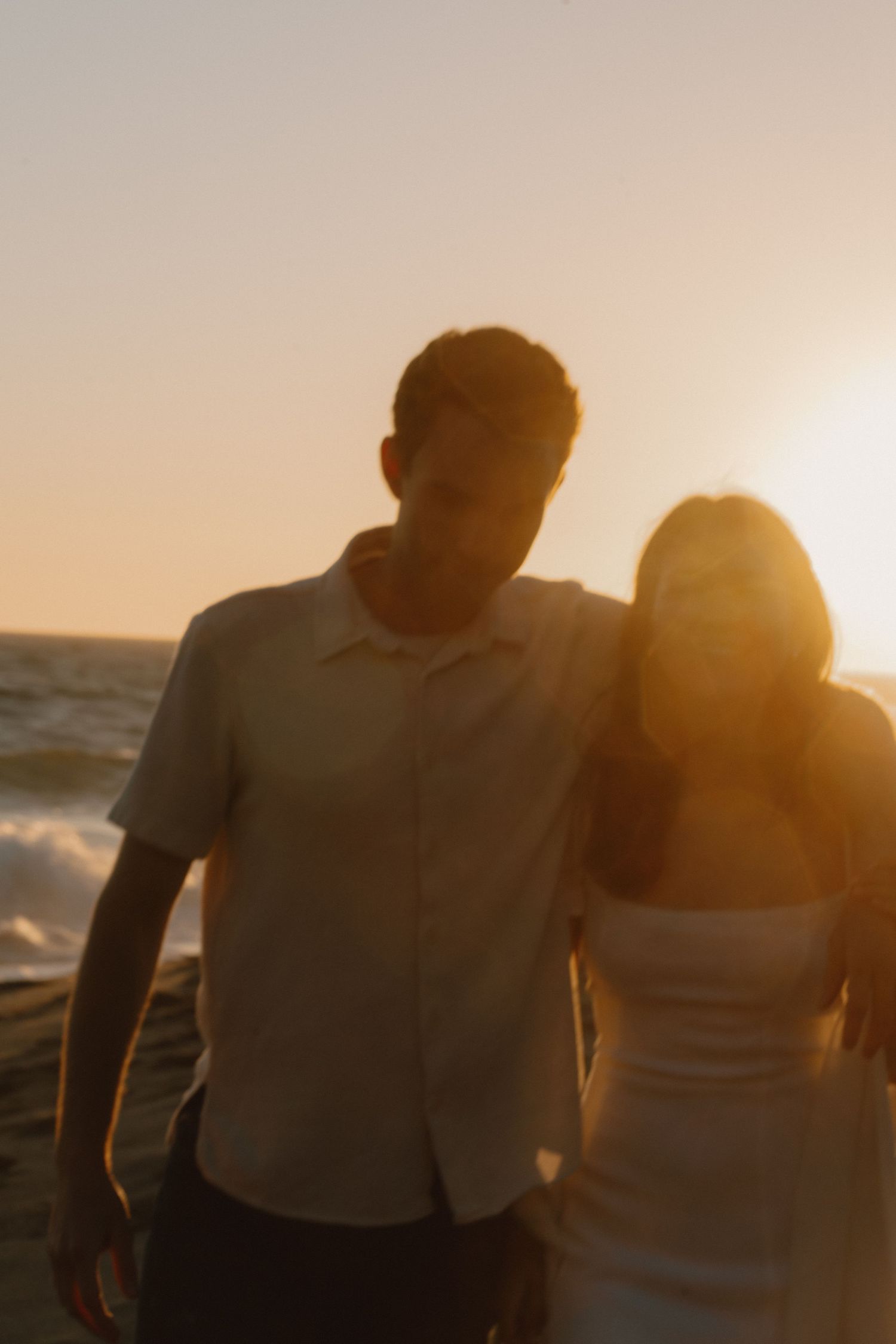 Couple walking on beach at golden sunset, waves crashing behind them in warm glowing light.