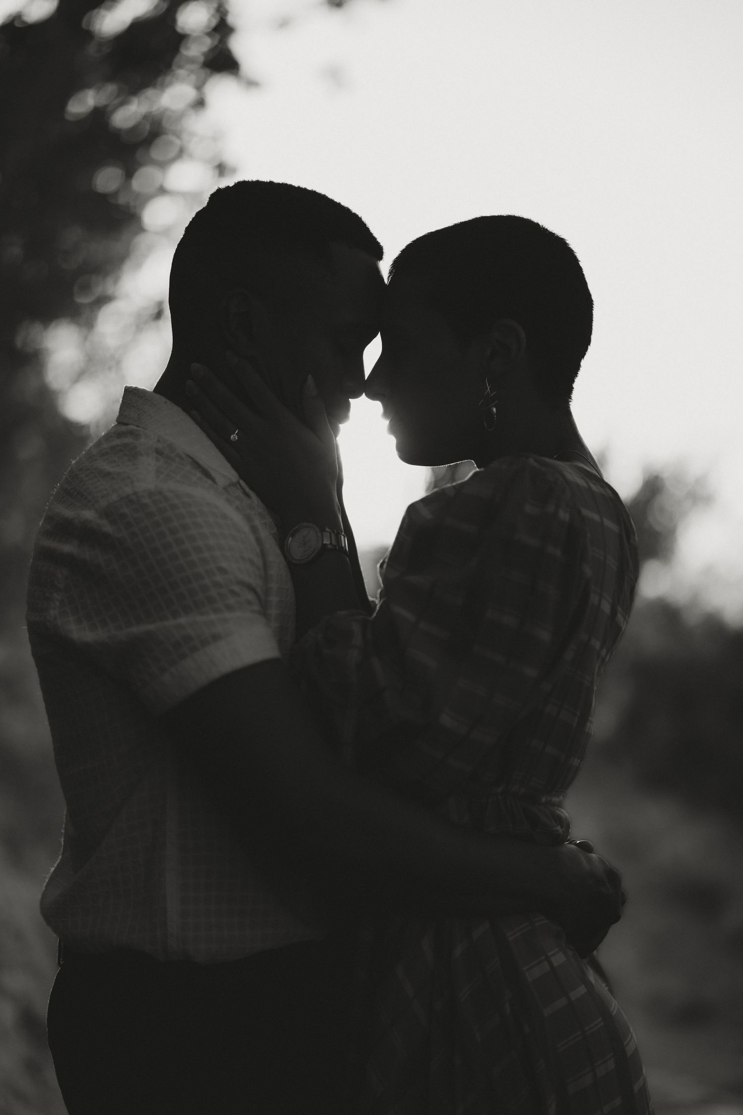 Black and white silhouette of a romantic couple embracing and touching foreheads outdoors among trees.