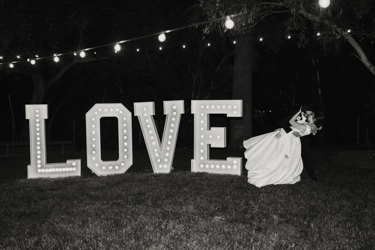 Black and white photo of illuminated LOVE marquee letters on grass under string lights at wedding reception.