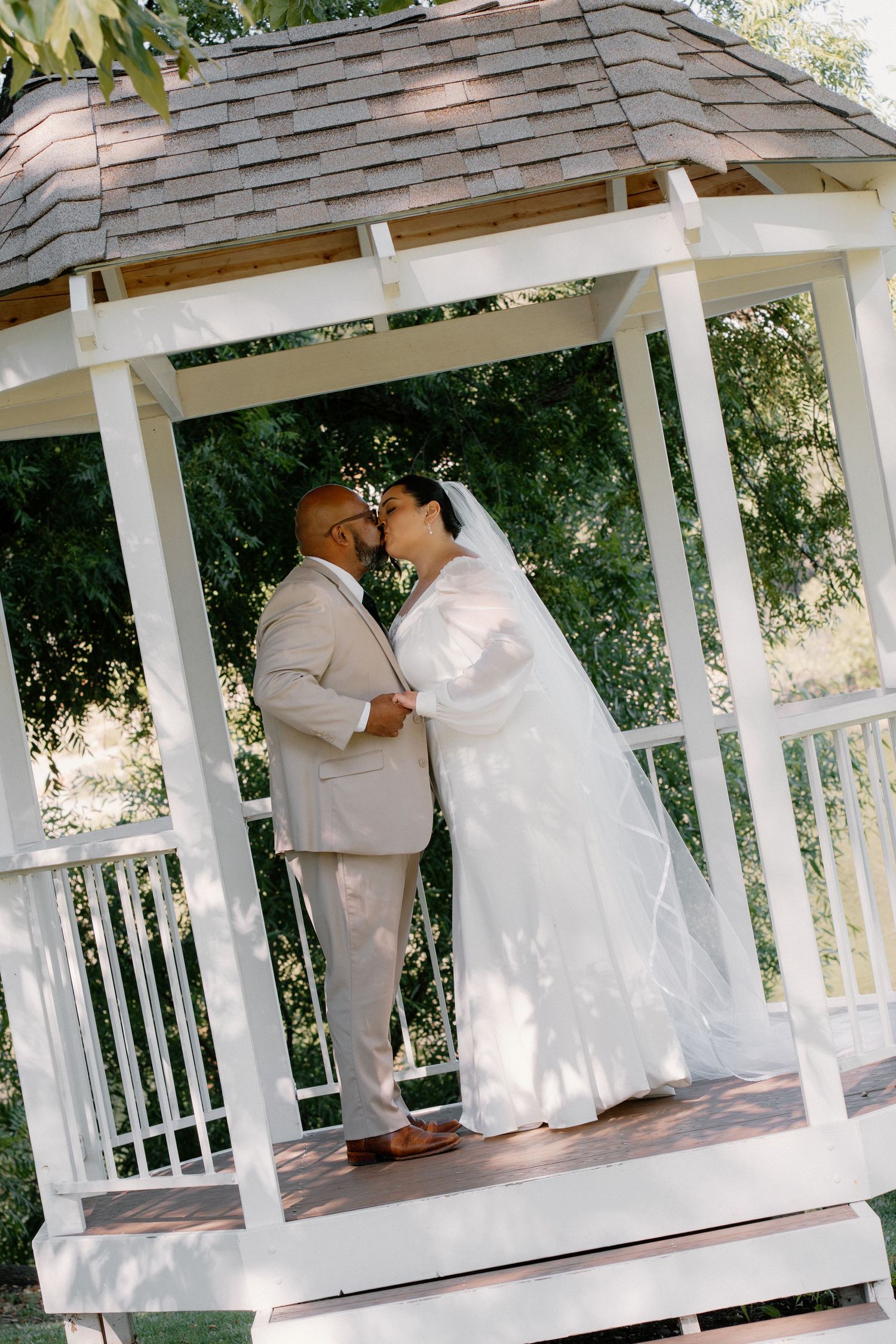 Bride and groom sharing a kiss inside a white wooden gazebo surrounded by lush green trees.