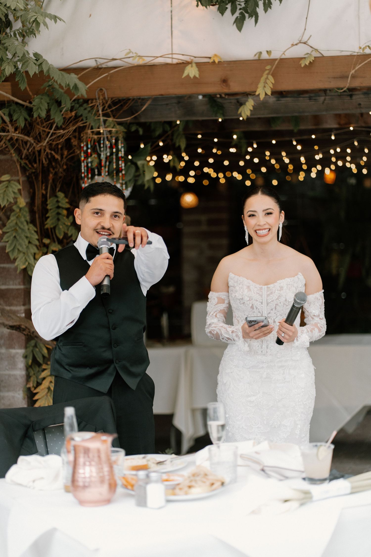 Bride and groom smiling while giving speeches with microphones at elegant outdoor wedding reception.