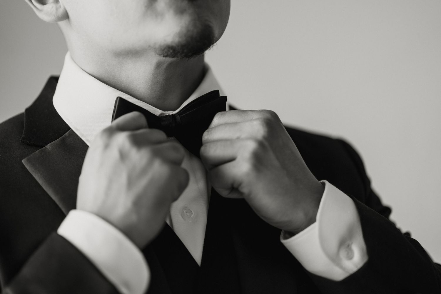 Close-up black and white photo of a groom adjusting his elegant black bow tie before the wedding ceremony.