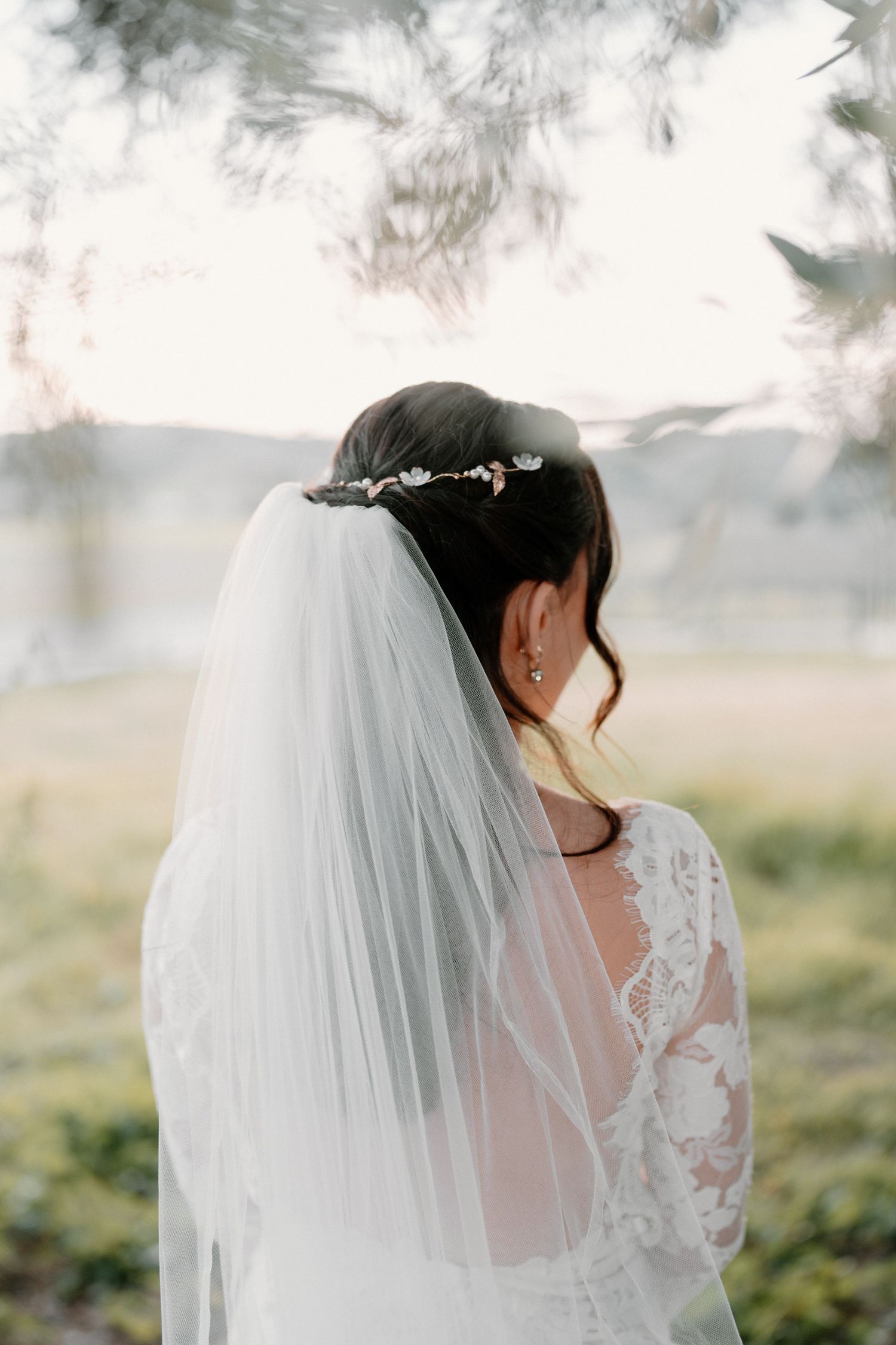 Back view of bride with dark hair in lace dress wearing floral hair comb and white veil outdoors.