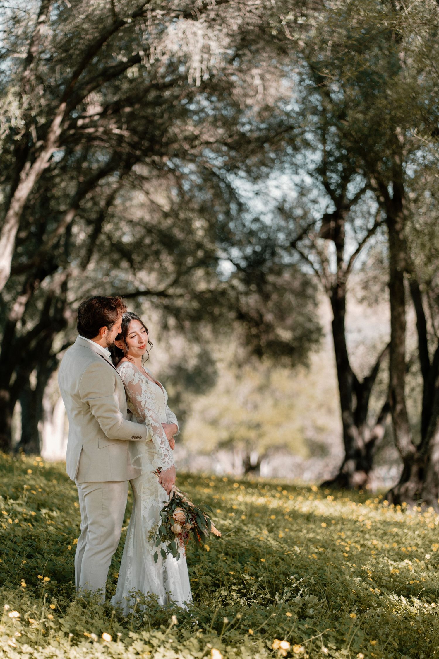 A bride and groom share a tender moment among olive trees on a sunny wedding day.