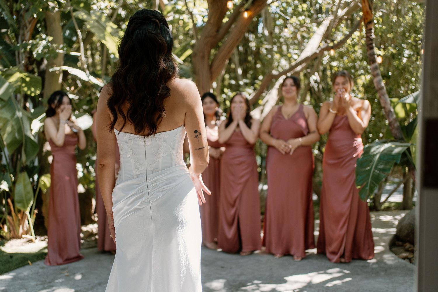 Bride in fitted white gown reveals dress to emotional bridesmaids in dusty rose dresses outdoors.