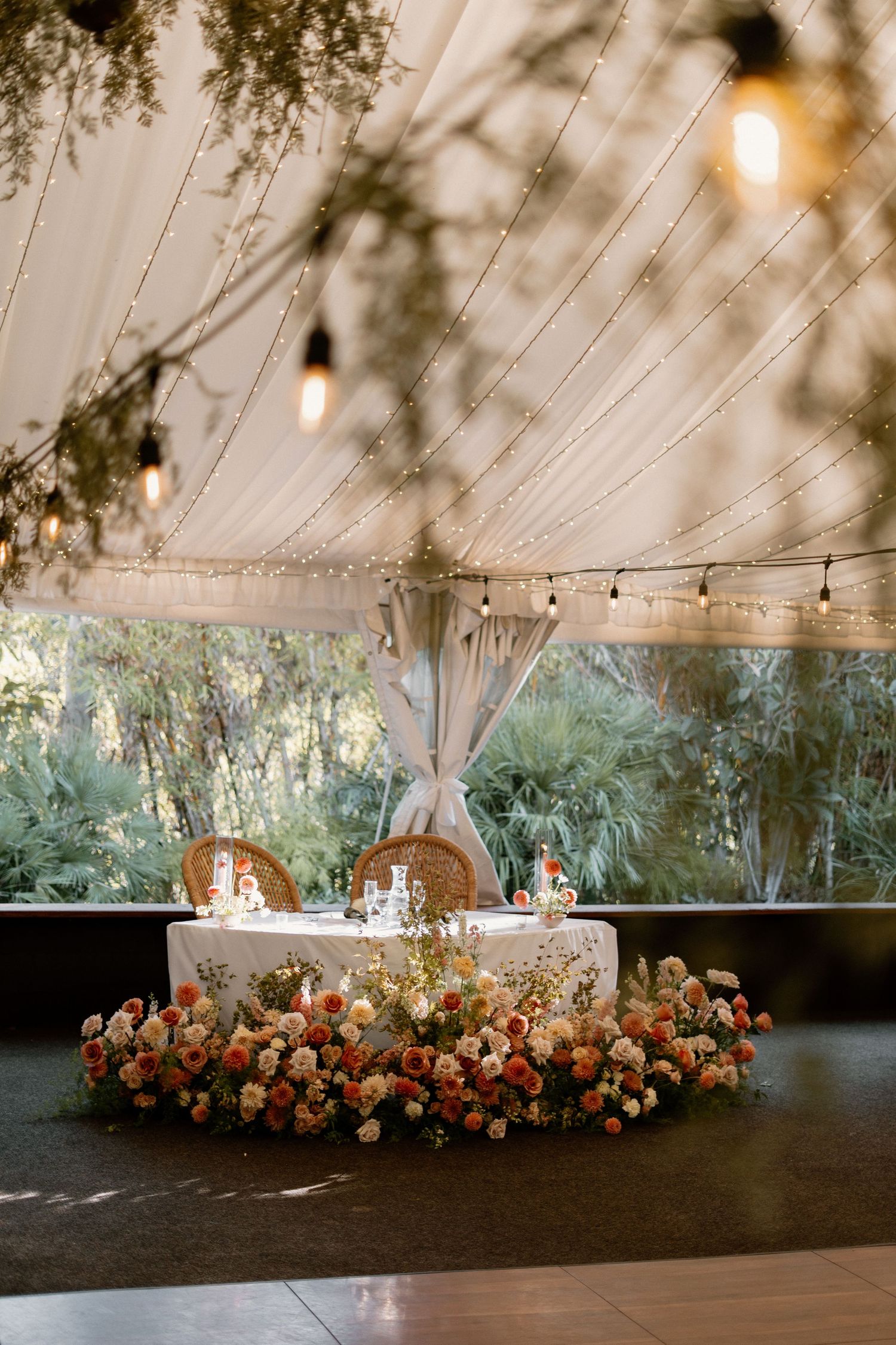 Elegant sweetheart table adorned with lush floral arrangement under string lights and draped tent ceiling.