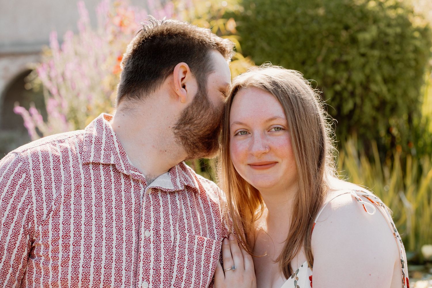 A man with a beard kisses a smiling woman on the cheek outdoors in golden sunlight near blooming lavender.