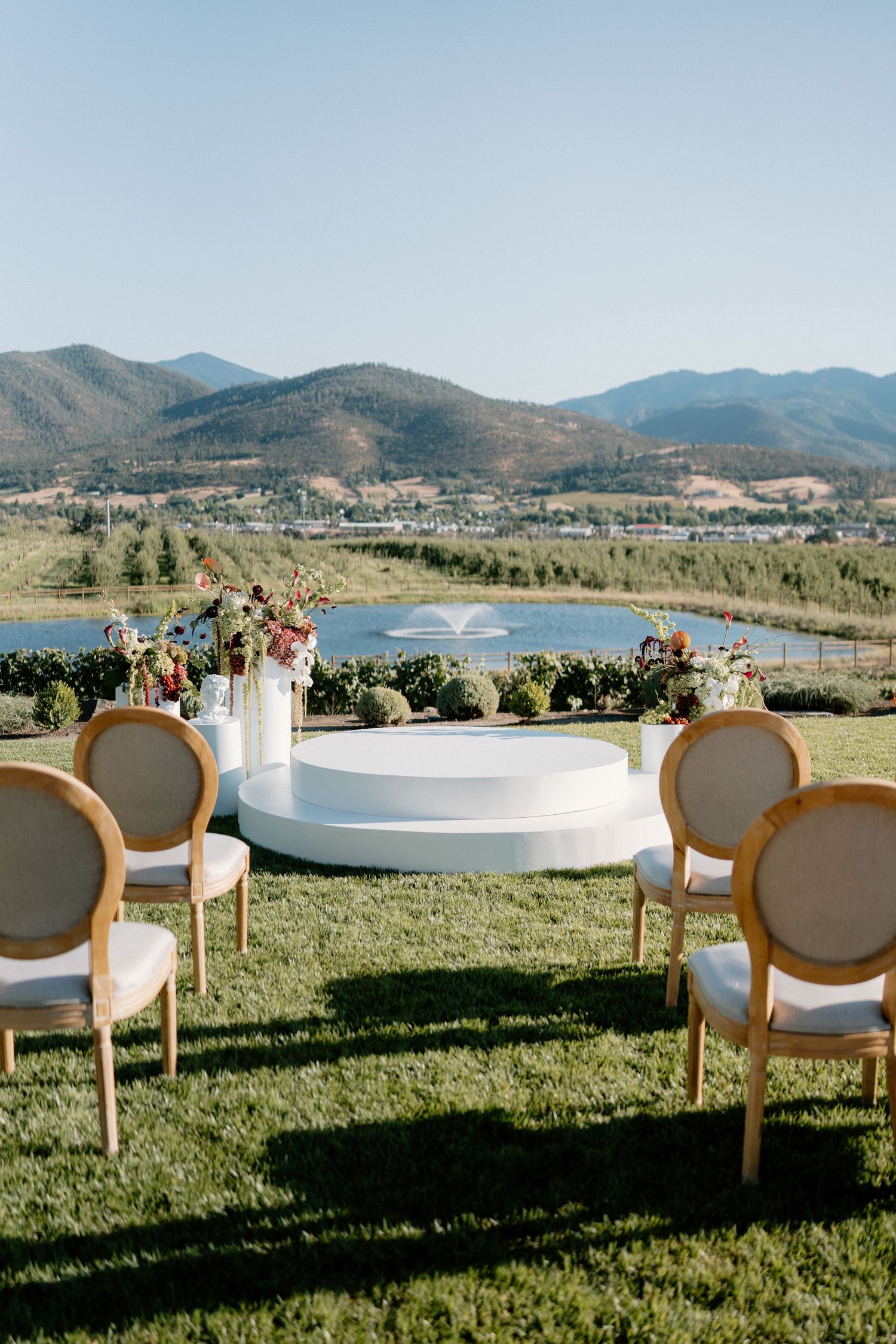 Outdoor wedding ceremony setup with white circular altar, gold chairs, lake, and mountain views.