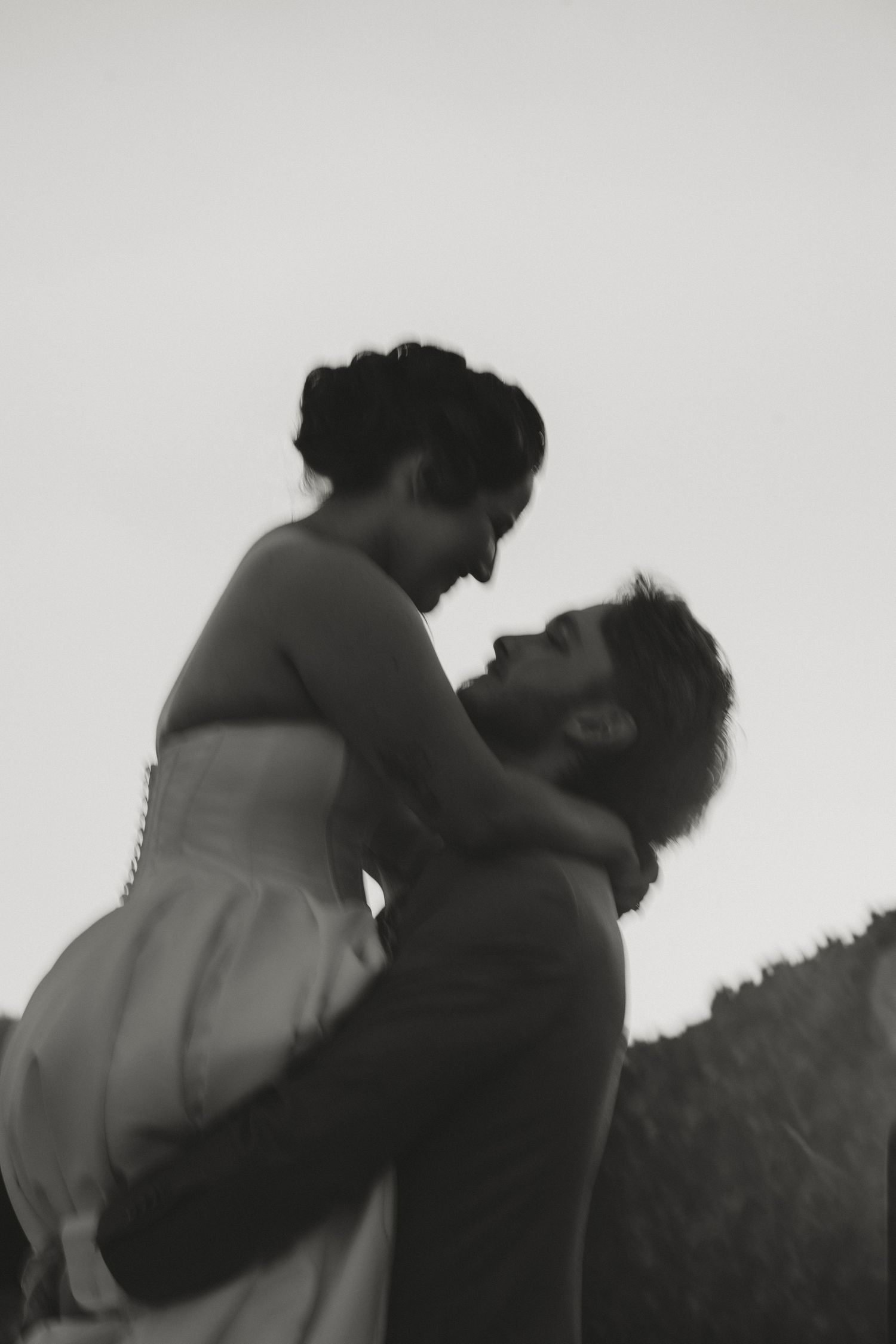 Black and white photo of groom lifting bride outdoors, sharing romantic moment against overcast sky.