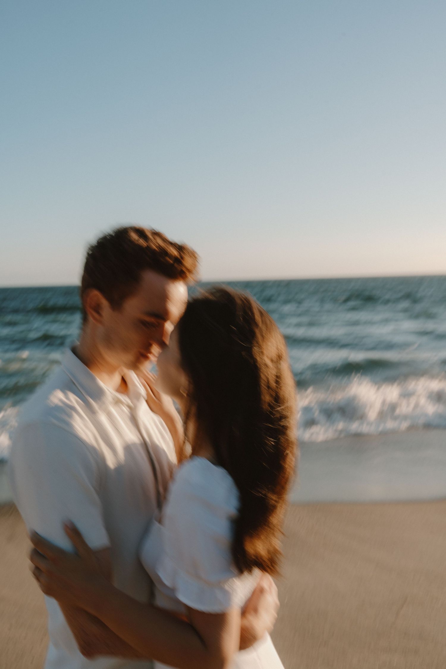 Blurred romantic couple embracing and almost kissing on sunny beach with ocean waves behind them.