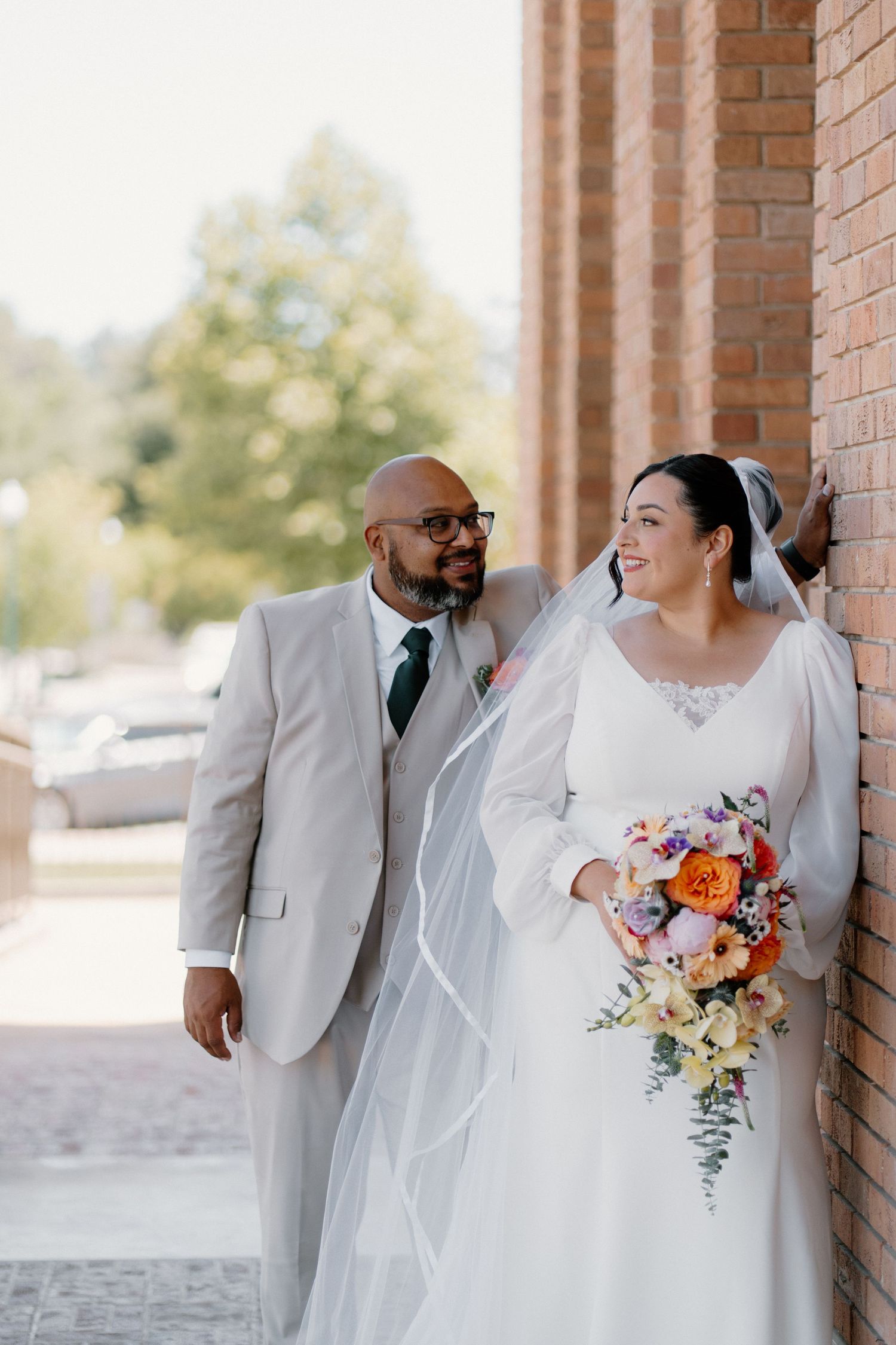Bride and groom pose beside a brick wall outdoors, she holds a colorful bouquet in a white gown with veil.