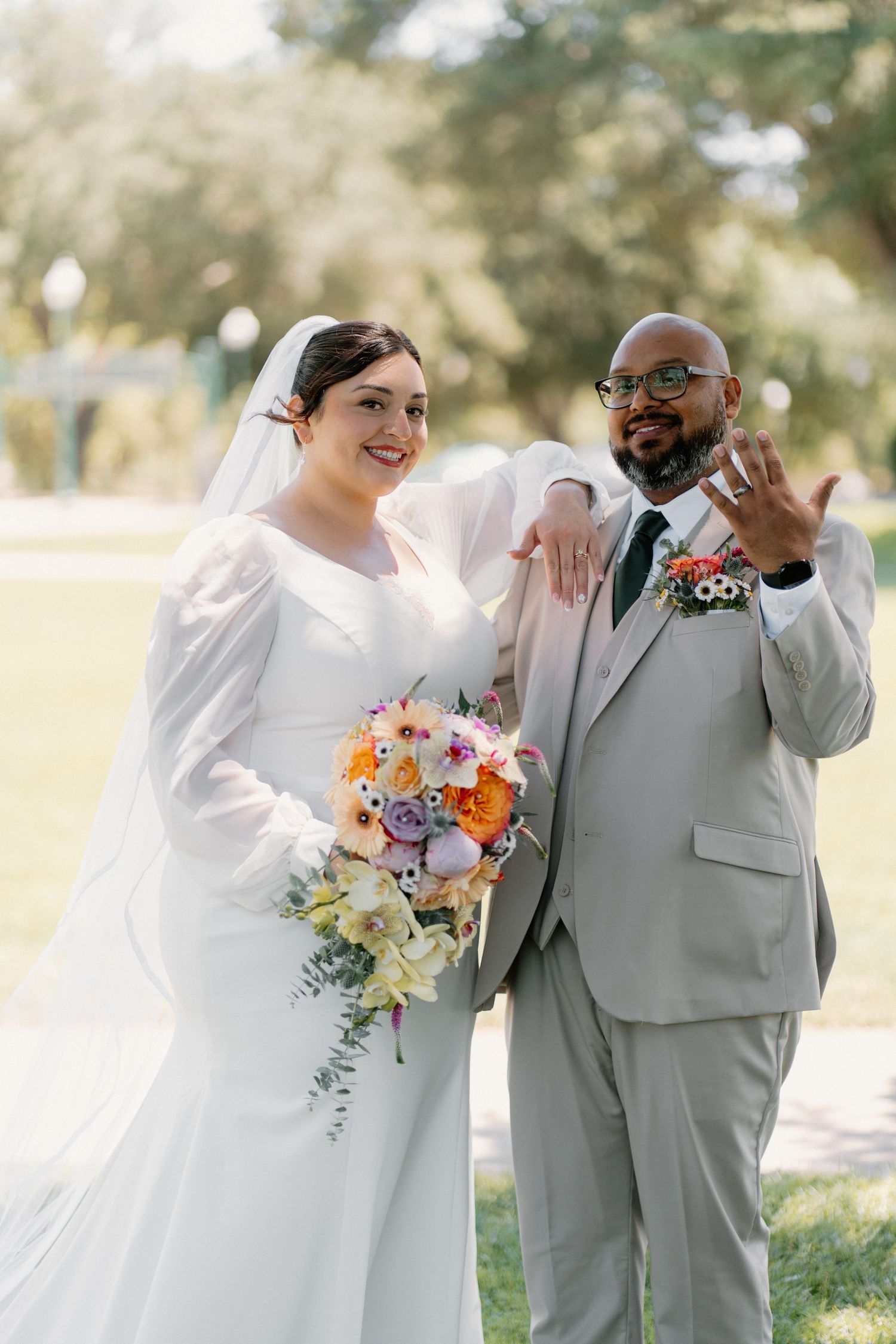 Bride and groom posing outdoors, showing wedding rings, bride holding colorful bouquet in white gown with veil.