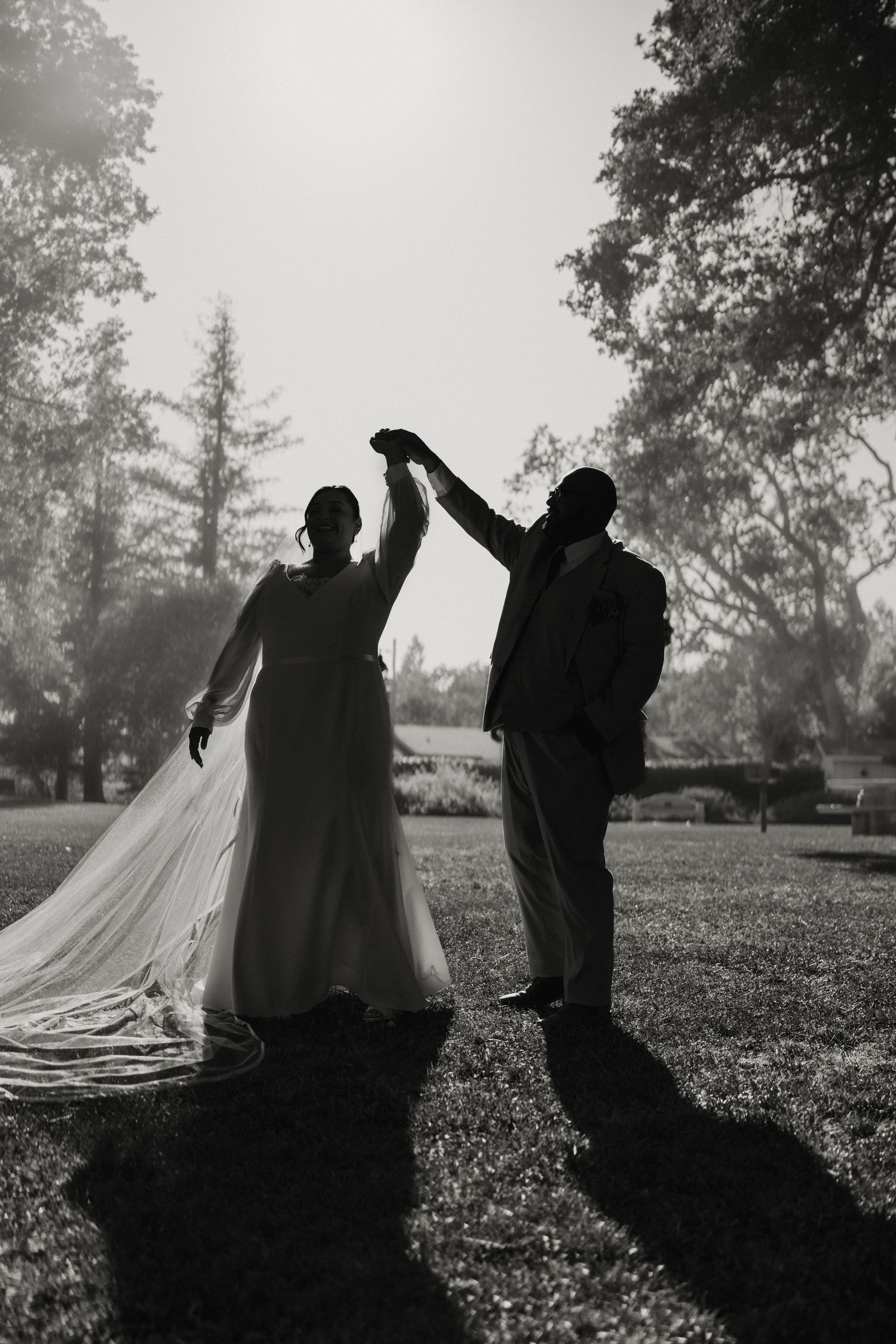 Black and white silhouette of bride and groom dancing outdoors, long veil flowing in sunlight.