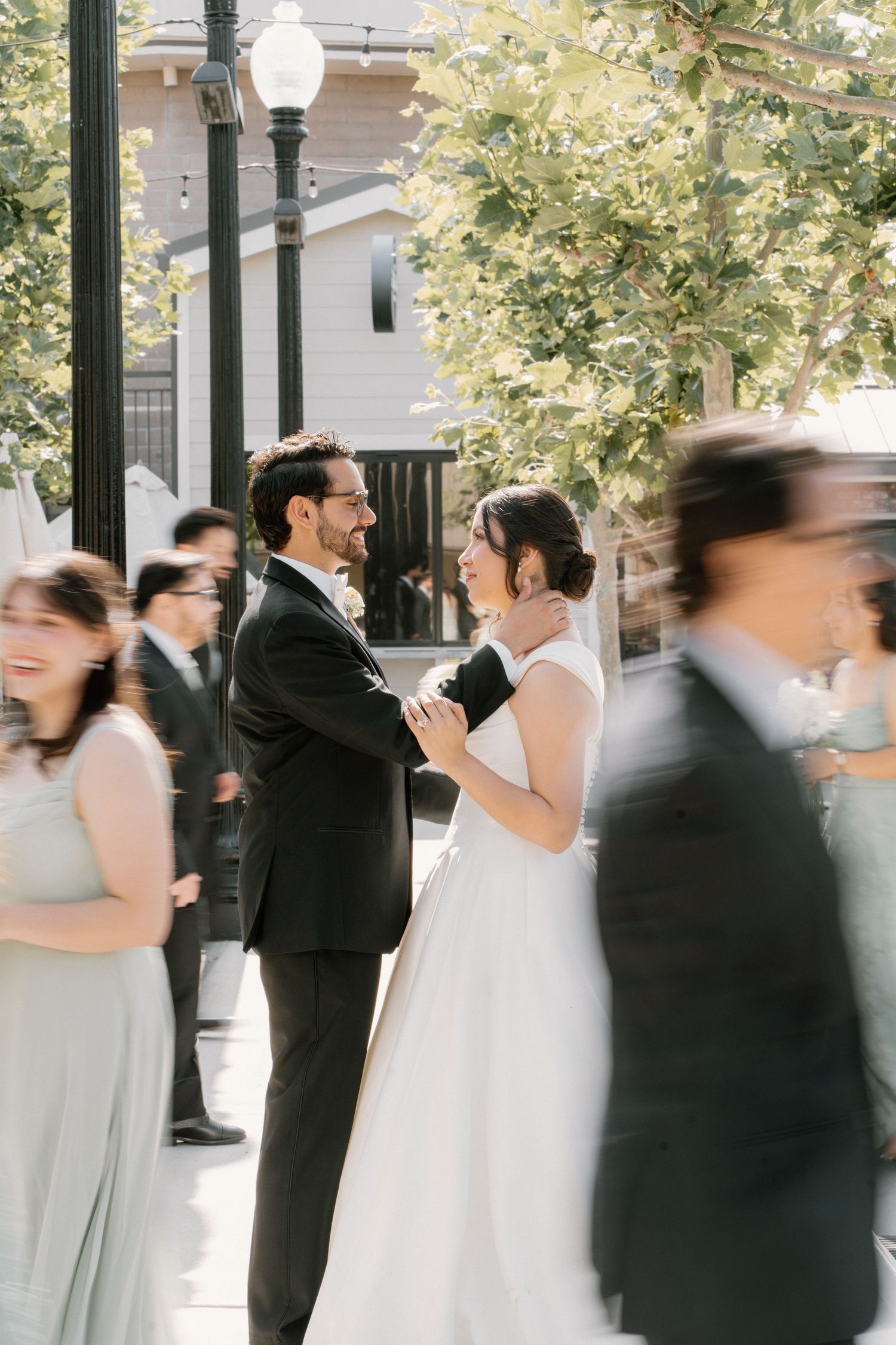 Bride and groom share a tender moment while wedding guests blur around them outdoors.