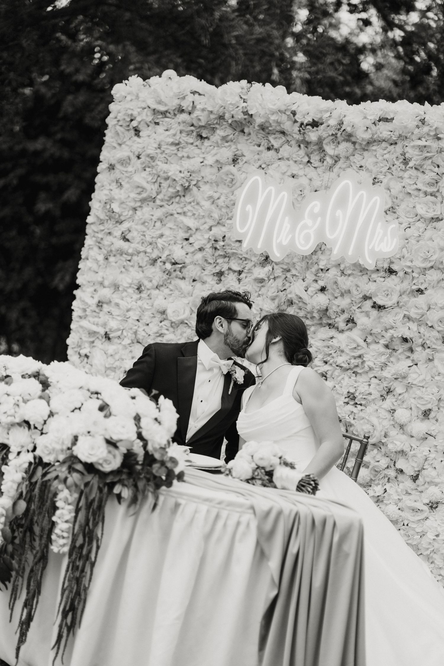 Bride and groom share a kiss at sweetheart table before floral wall backdrop with Mr & Mrs neon sign.
