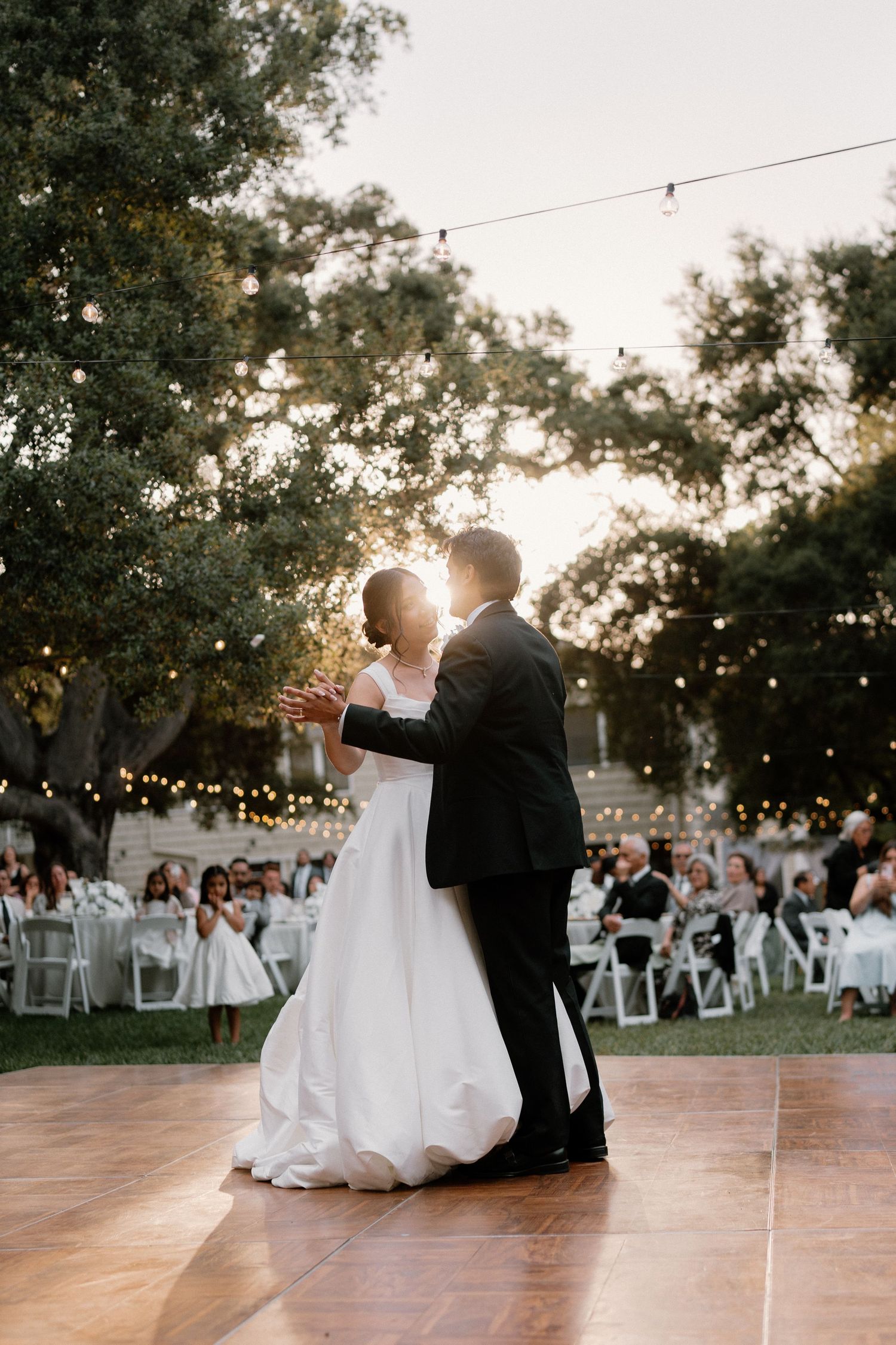 Bride and groom share romantic first dance under string lights at golden hour outdoor wedding reception.