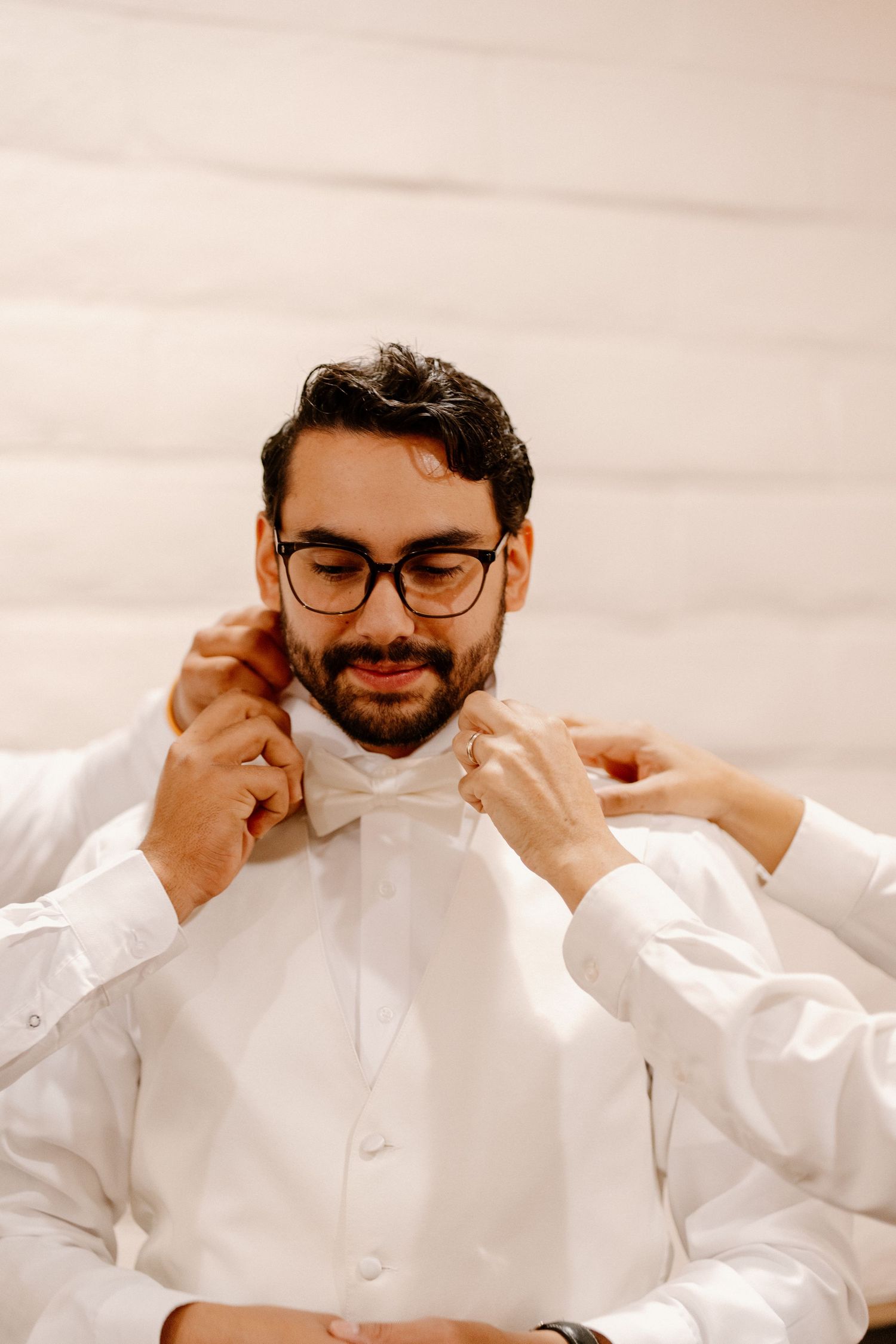 Groom in white shirt and glasses has white bow tie adjusted by hands during wedding day preparations.