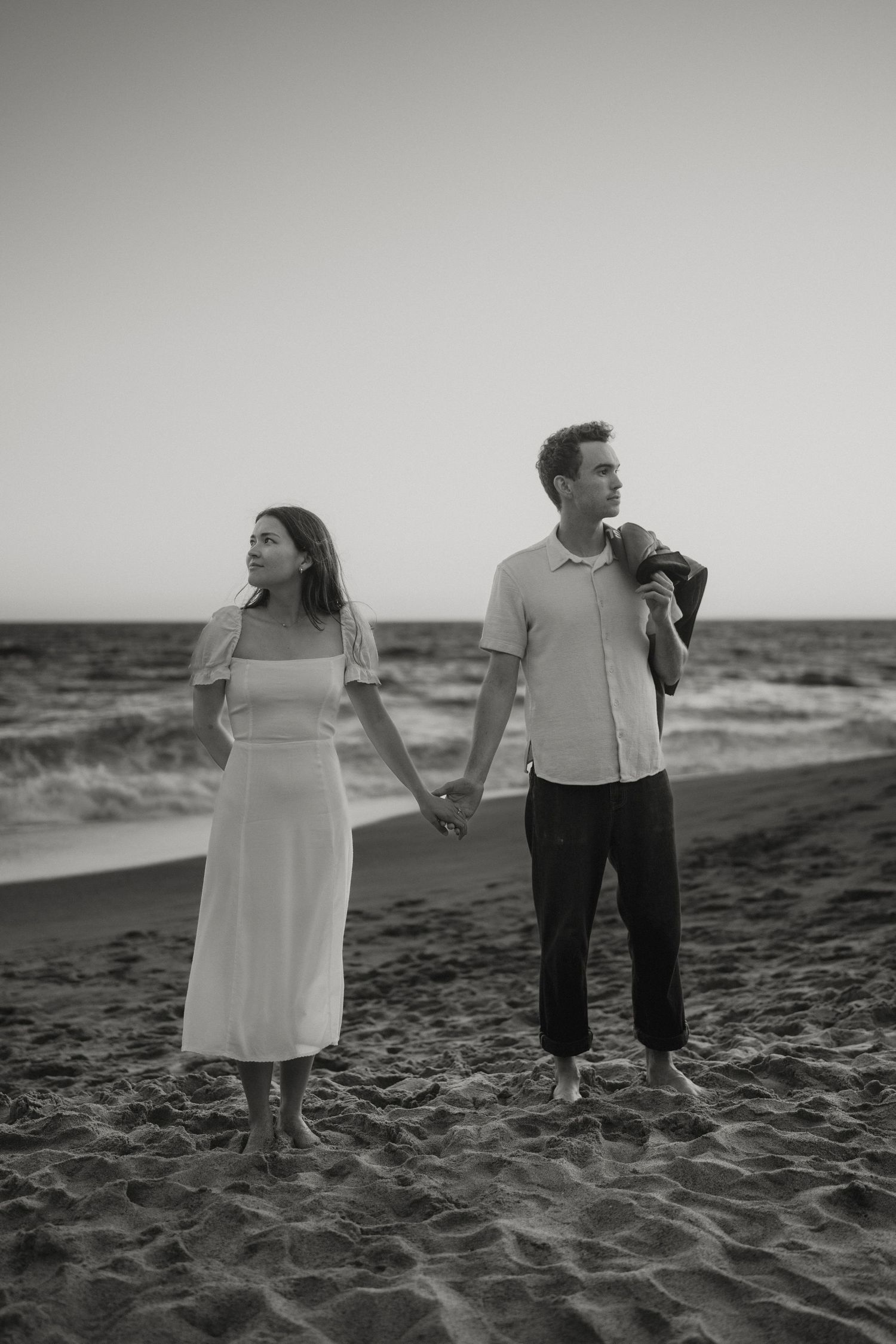 Black and white photo of a couple holding hands on a sandy beach at dusk, woman in white dress, man in light shirt.