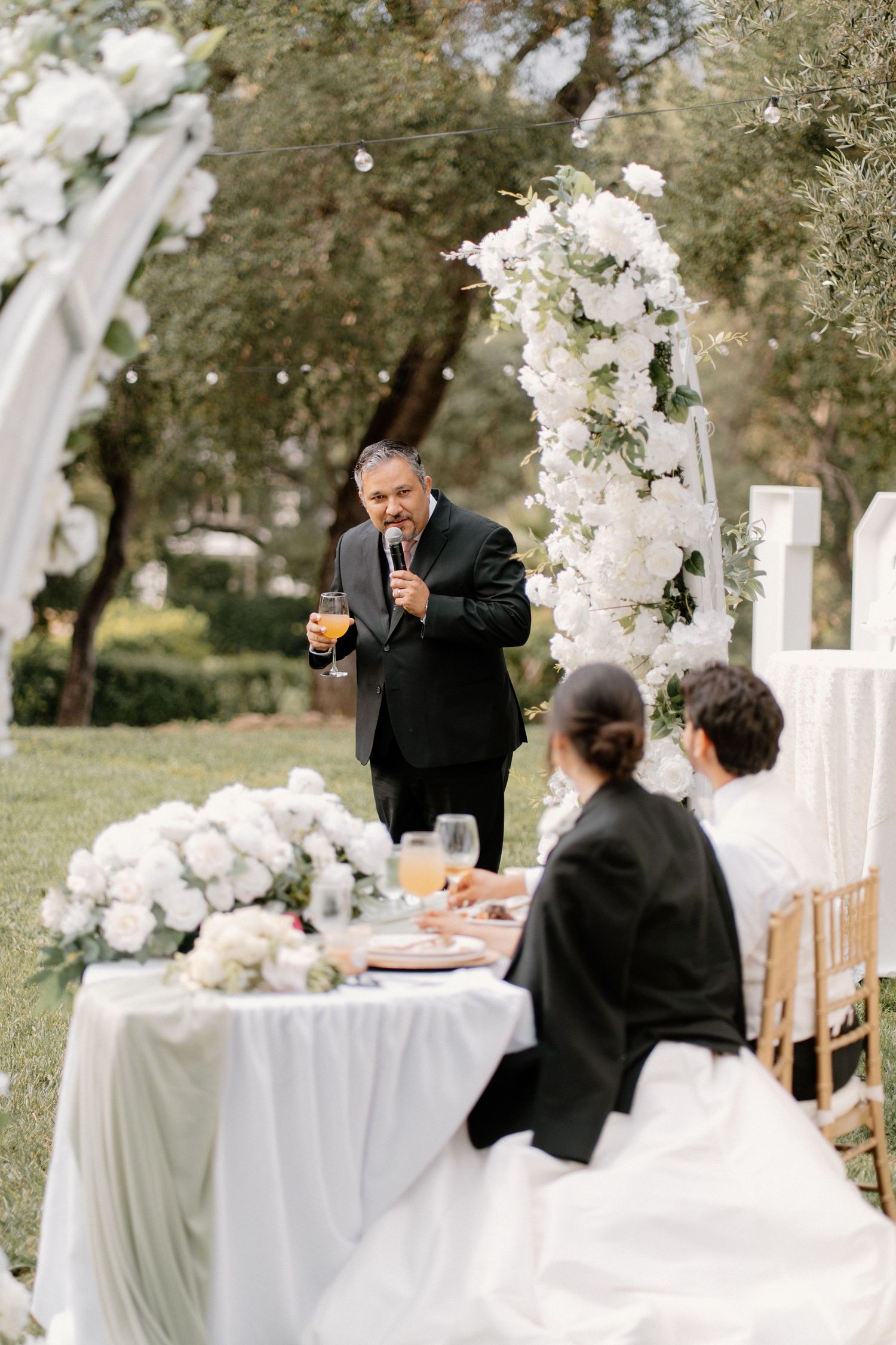 Father of bride gives heartfelt toast at outdoor wedding reception with white floral arch.