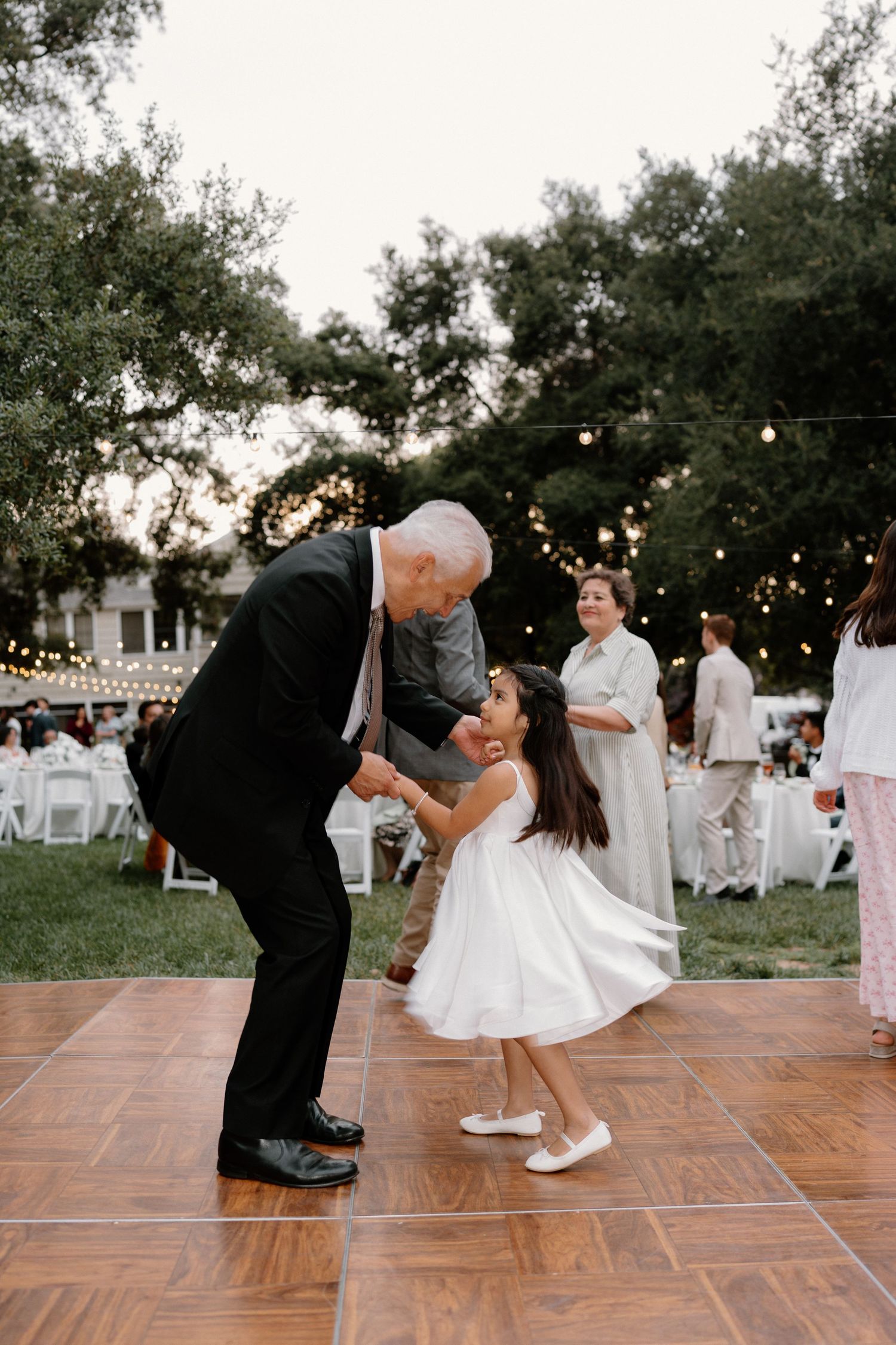 Elderly man in suit joyfully dances with young flower girl in white dress at outdoor wedding reception.