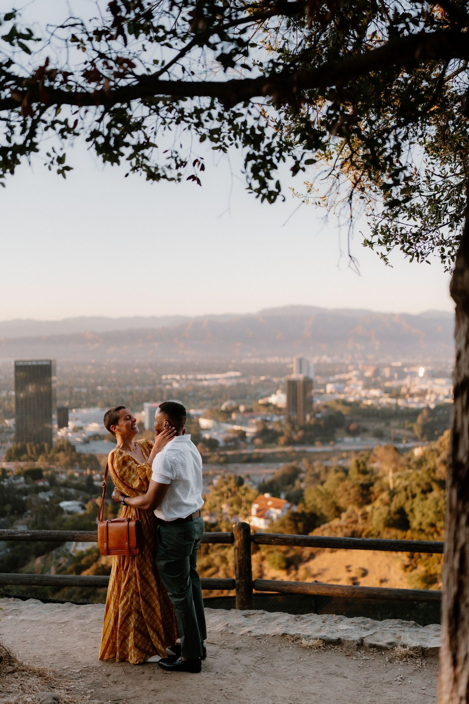 A couple shares a romantic moment under a tree at a scenic Los Angeles hillside overlook at sunset.