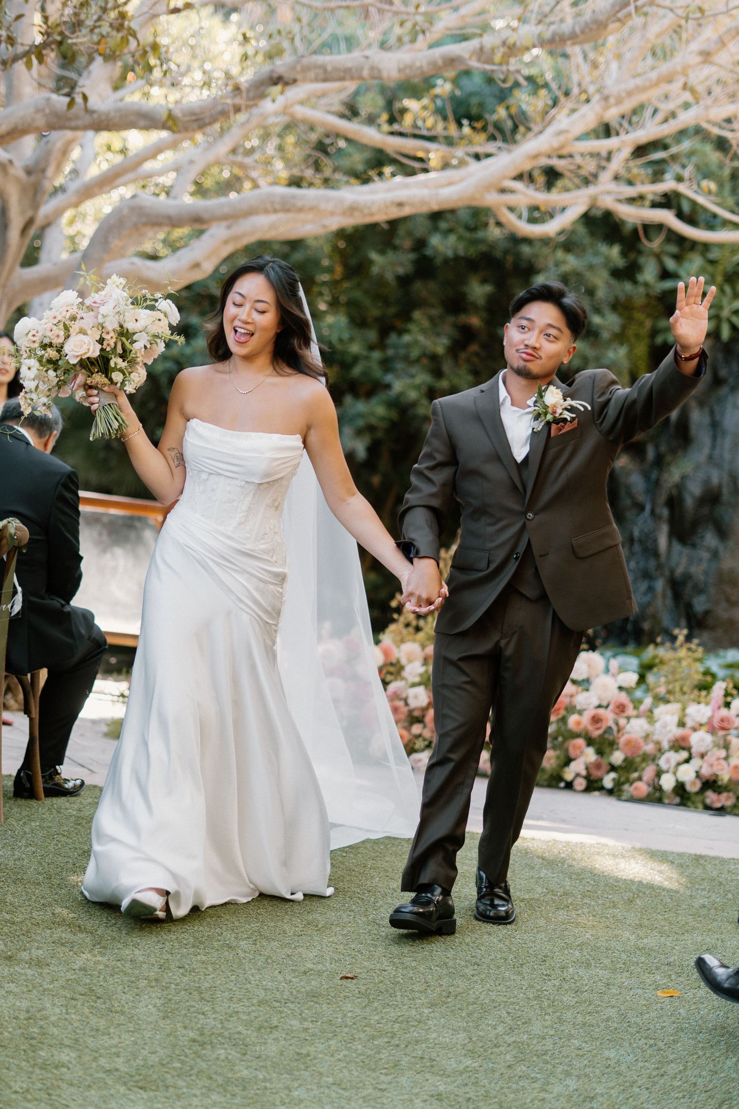 Newlyweds walk joyfully down the aisle at an outdoor garden wedding ceremony, bride holding bouquet.