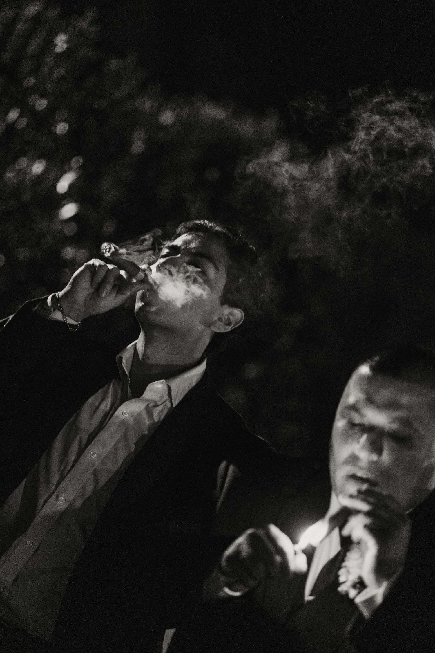 Two men smoking cigars outdoors at night in black and white, exhaling smoke dramatically.
