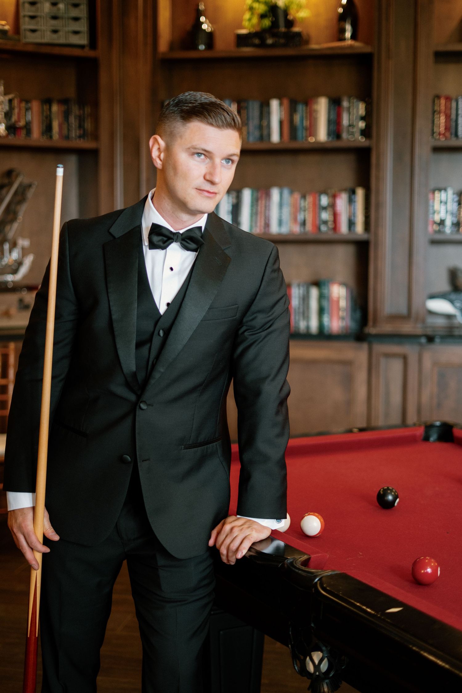 Elegant man in black tuxedo with bow tie holding a billiard cue beside a red pool table in a sophisticated library.