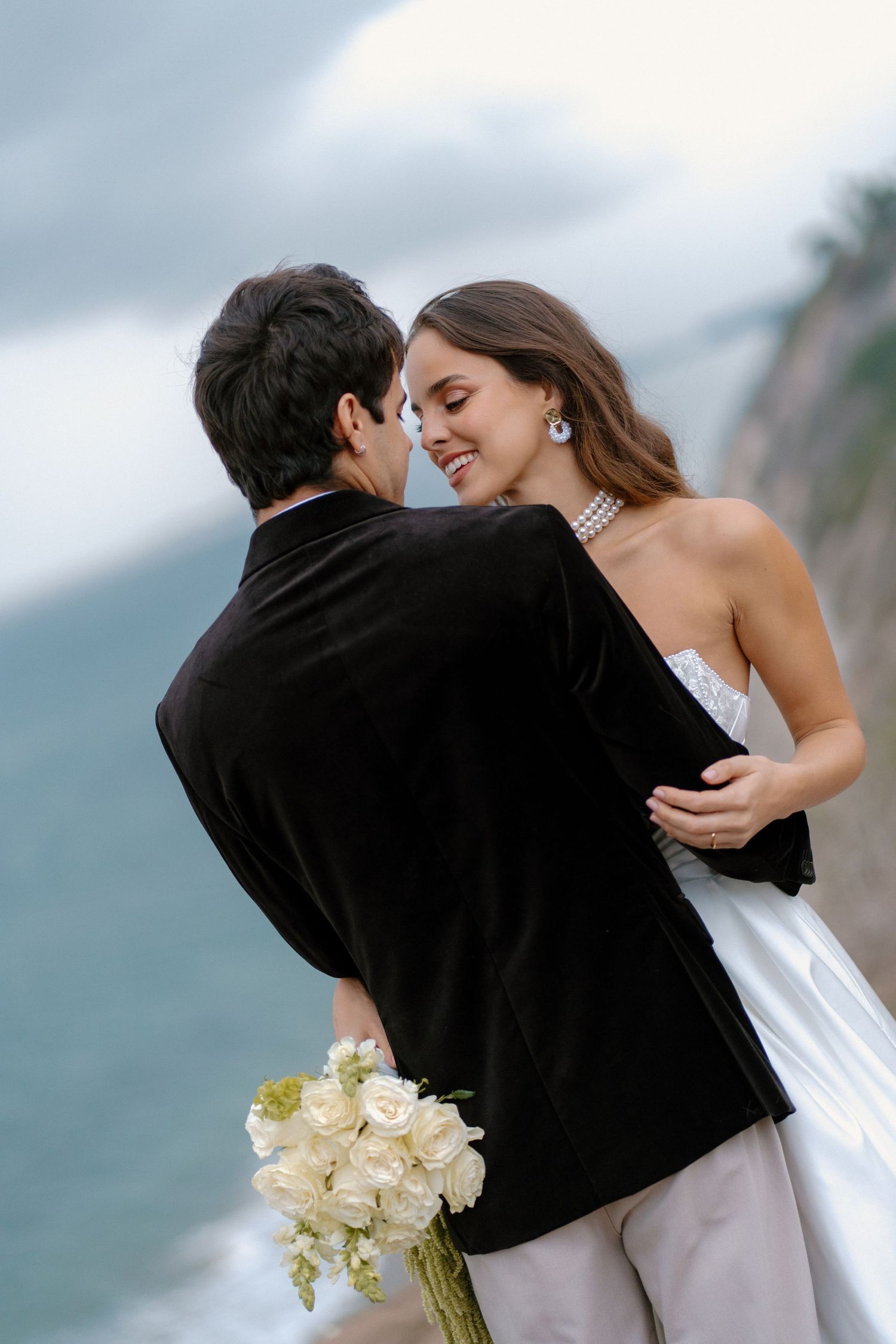 Romantic couple nearly kissing outdoors, bride in strapless white gown smiles as groom holds white rose bouquet by cliff.