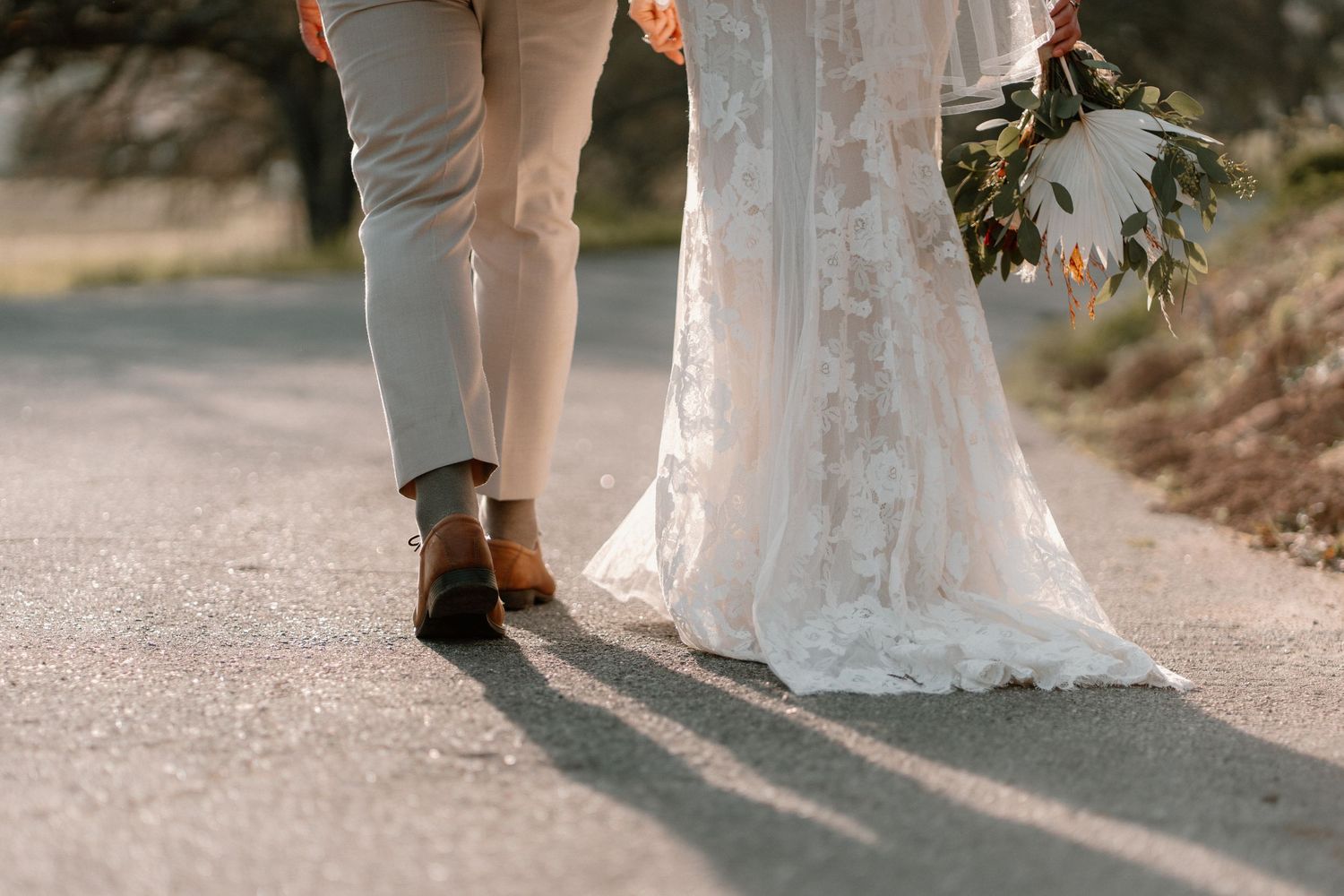 Close-up of bride in lace gown and groom in tan suit walking on a road holding a wedding bouquet.