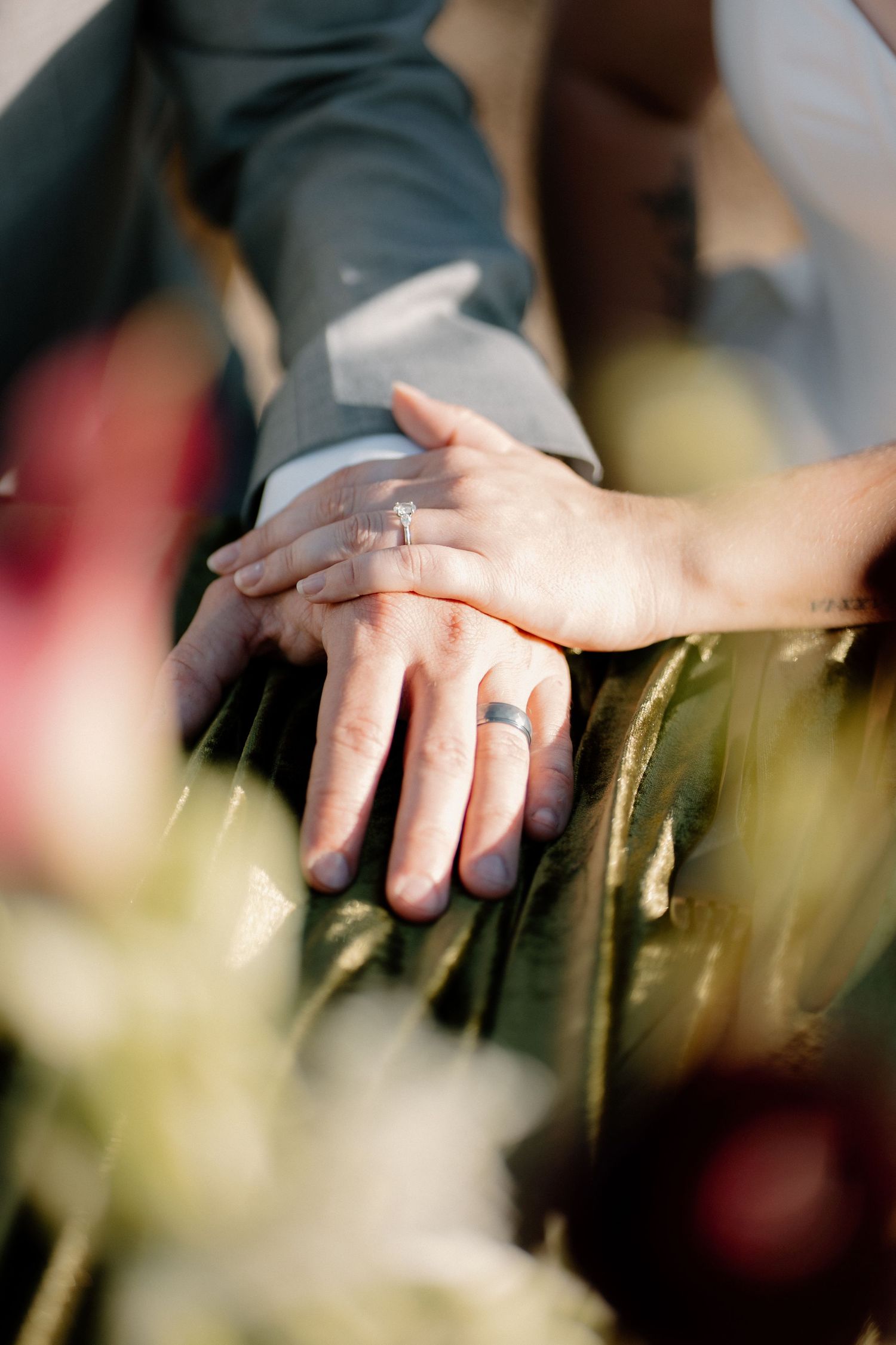 Close-up of newlyweds hands showing engagement ring and wedding bands resting on green foliage.