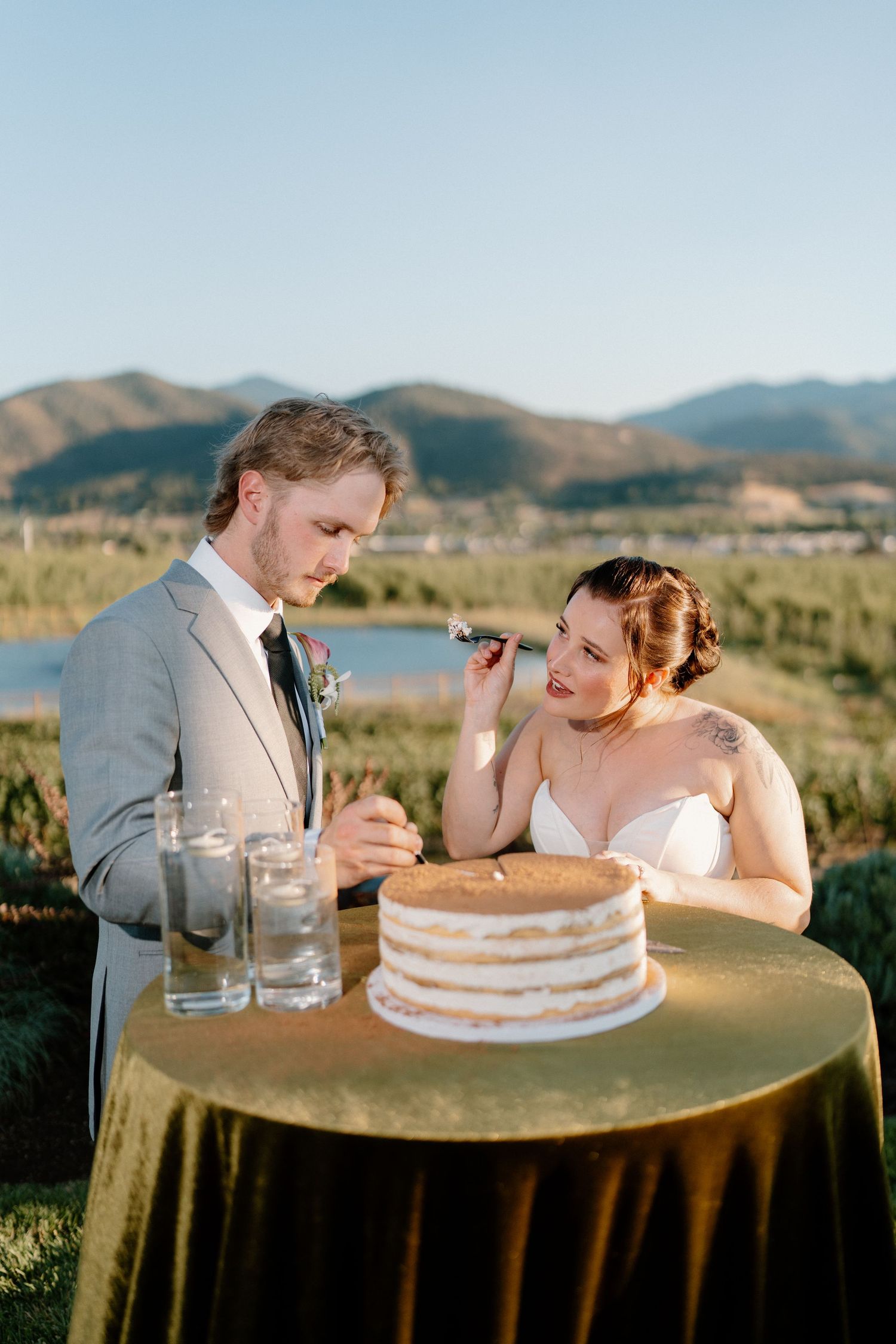Bride and groom cutting naked wedding cake outdoors with mountain lake backdrop at sunset reception.