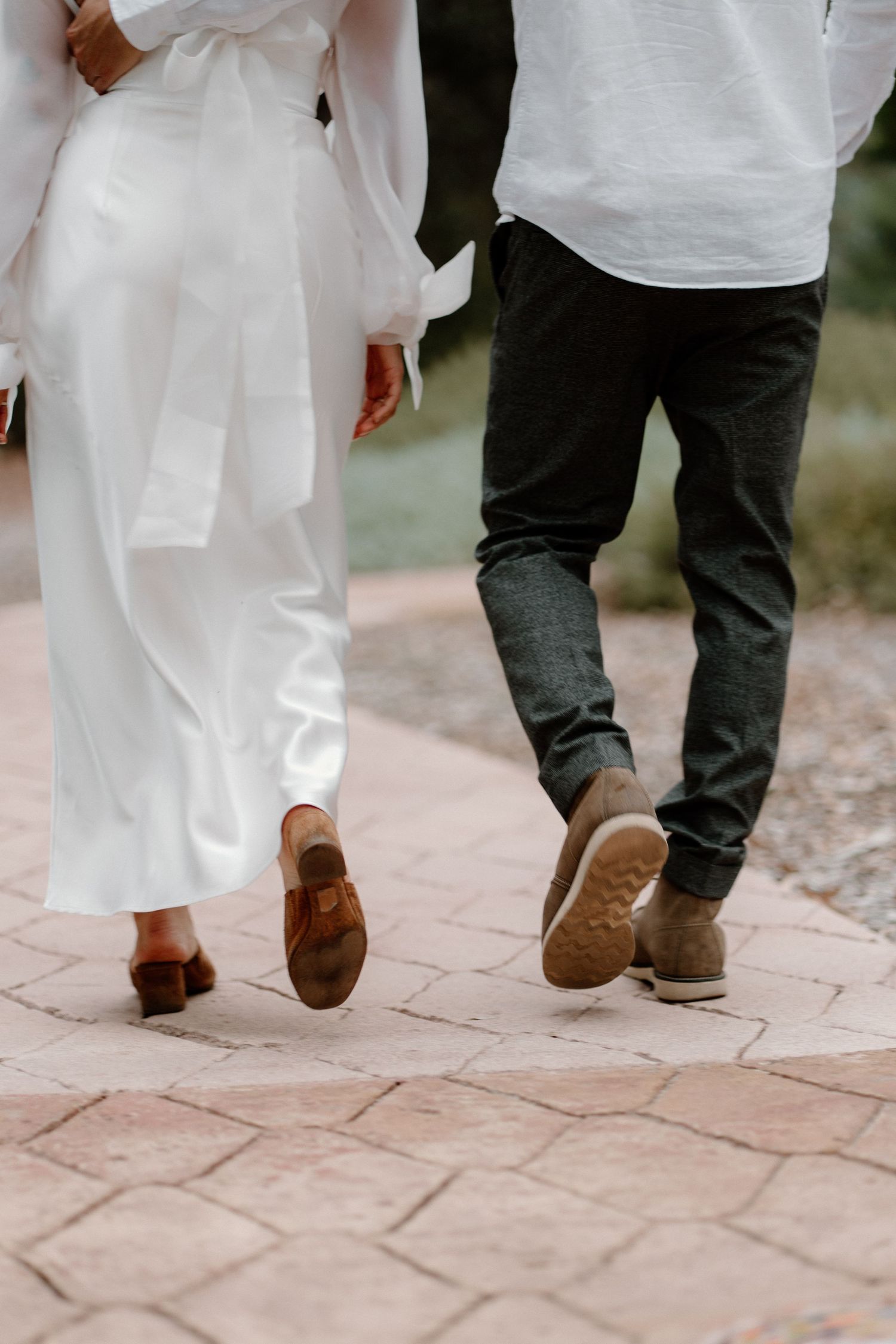 Close-up of couple walking on brick path, bride in white dress with brown clogs and groom in grey jeans.