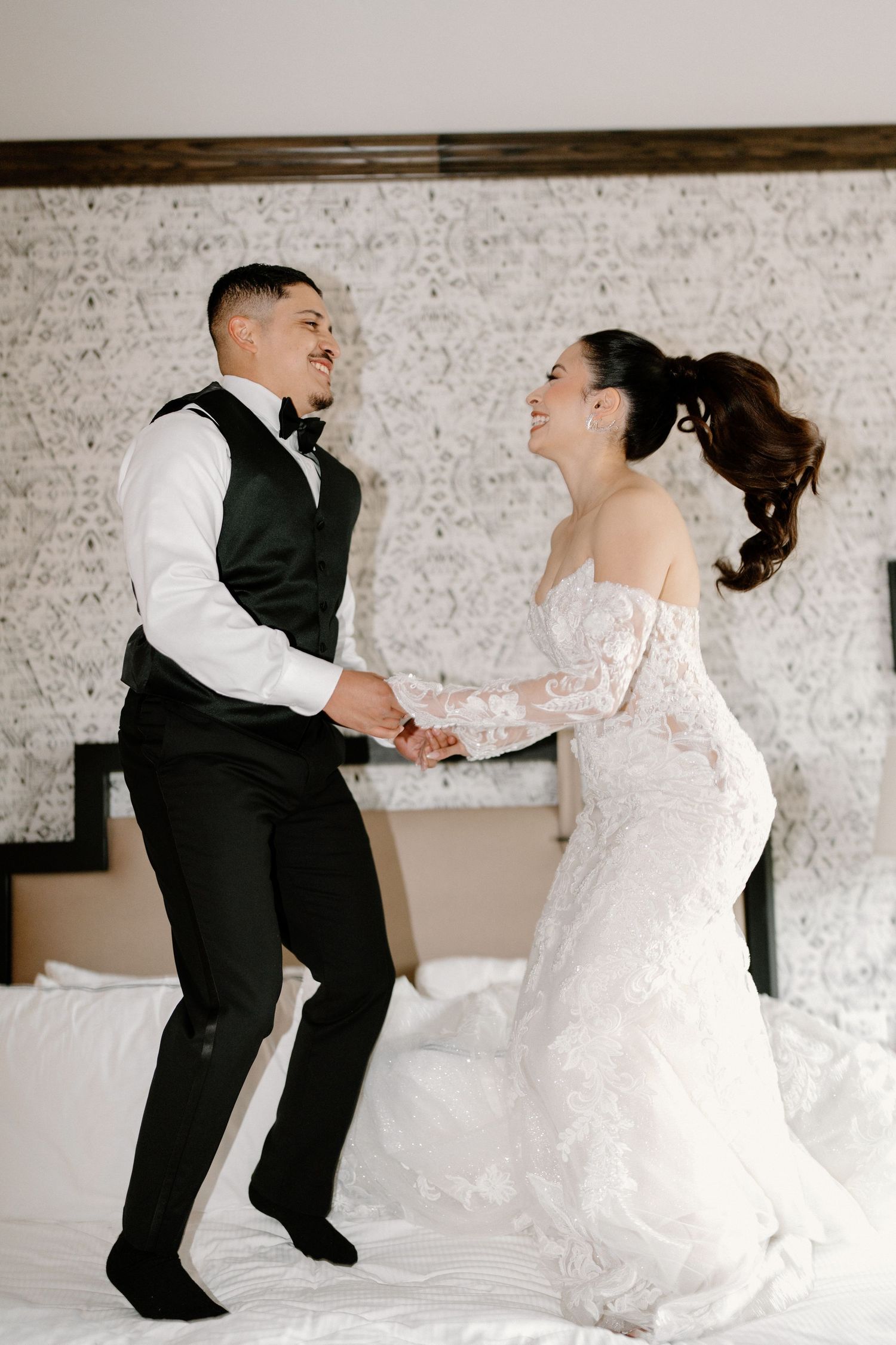 Joyful bride and groom hold hands while jumping on a hotel bed in their wedding attire.