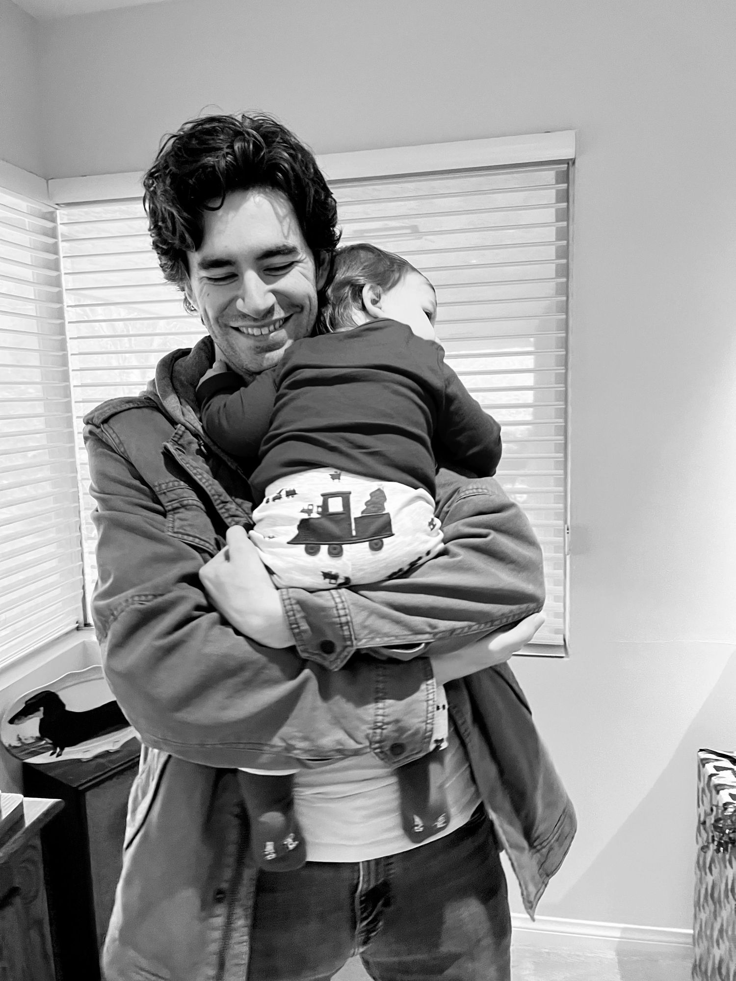 Black and white photo of a smiling man holding a baby close to his chest indoors.