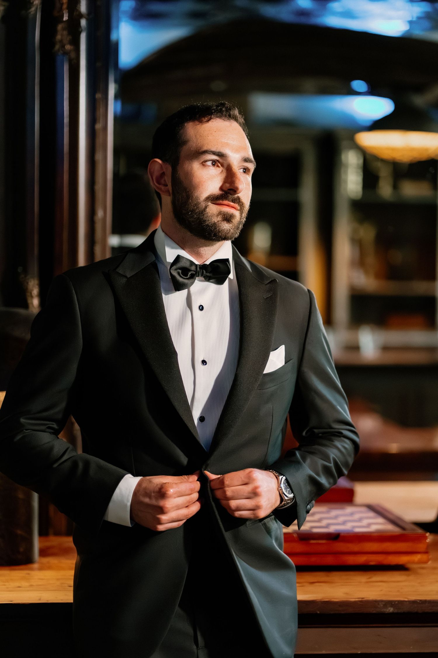 Dapper groom in black tuxedo with bow tie buttoning jacket in dimly lit elegant venue.