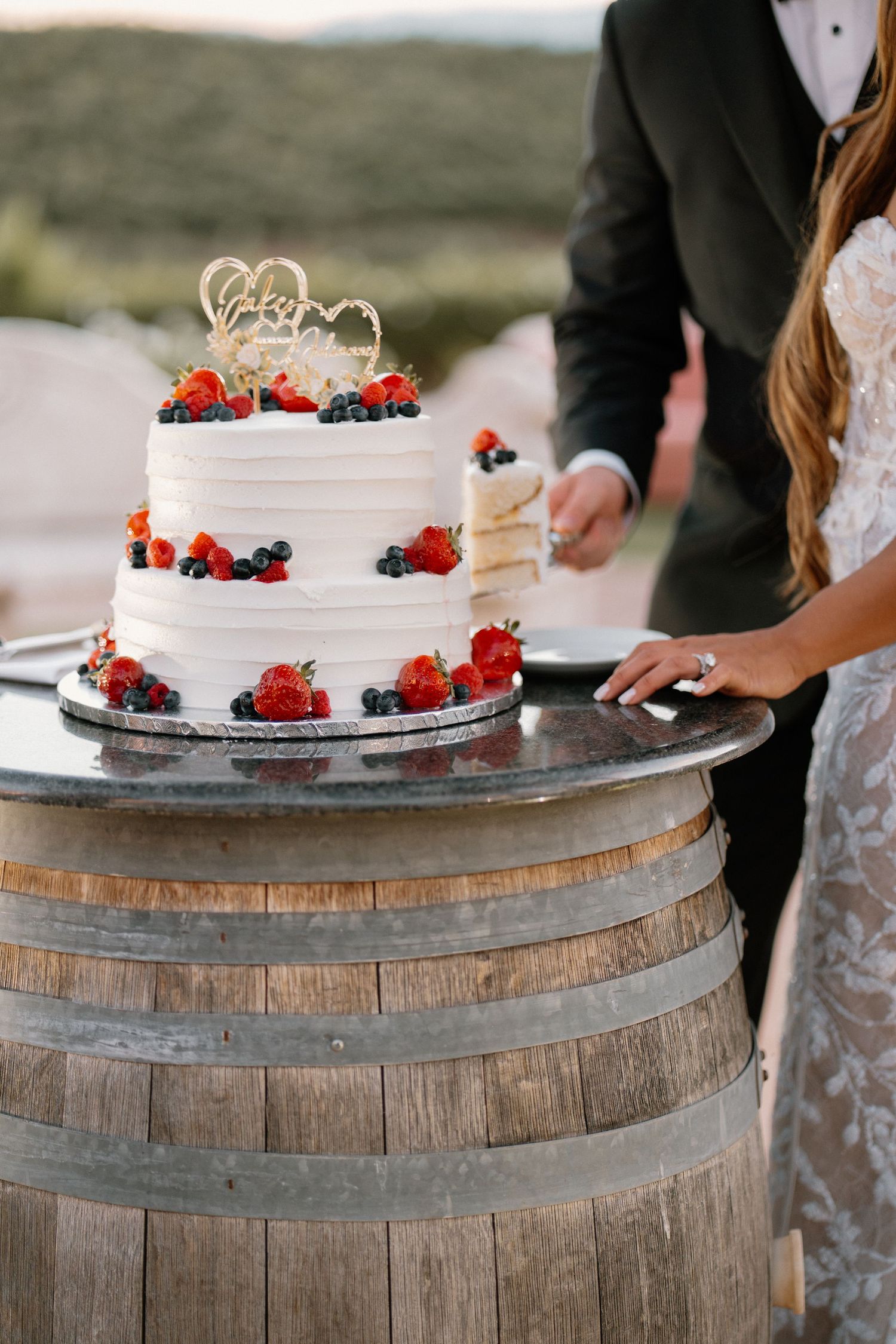 Couple cuts two-tier white wedding cake decorated with fresh berries displayed on rustic wooden barrel.