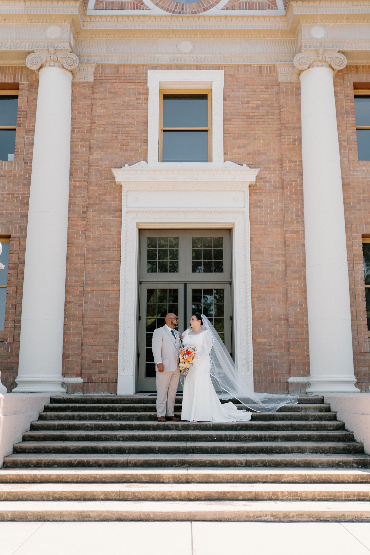 Bride and groom posing on steps of a brick building with white columns on their wedding day.