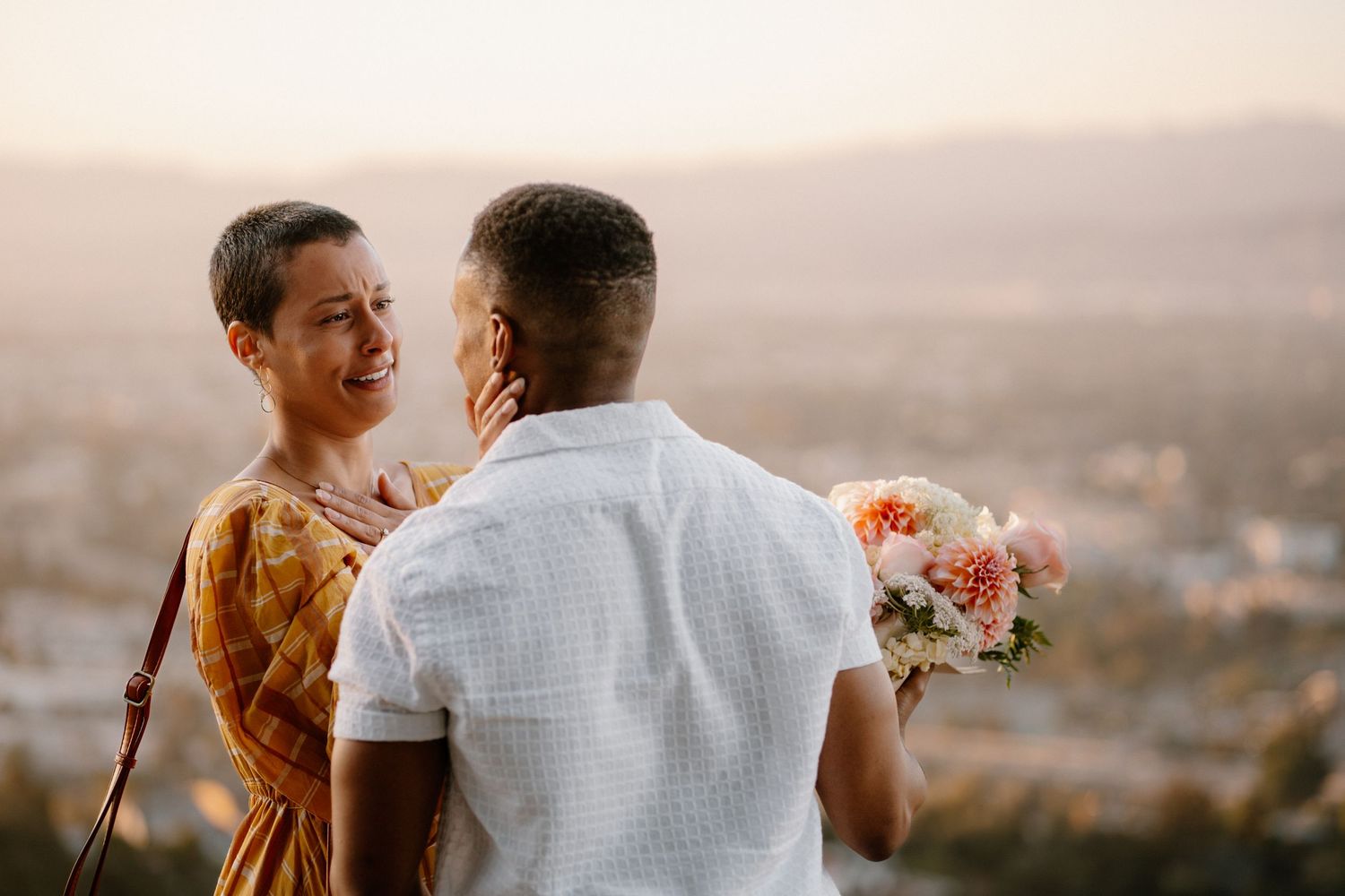 A man surprises a smiling woman with a bouquet of flowers at a scenic overlook during golden hour.